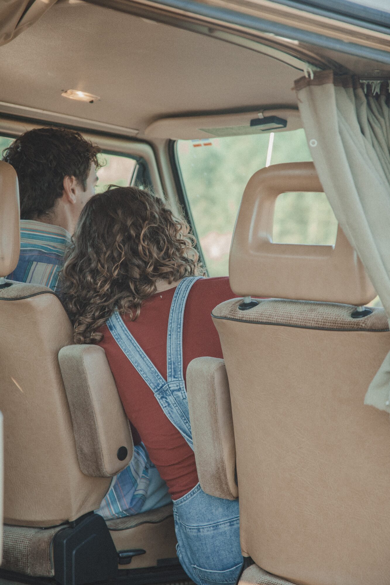 Two people sitting in a vehicle, viewed from behind, with a curtain partially visible on the right side.
