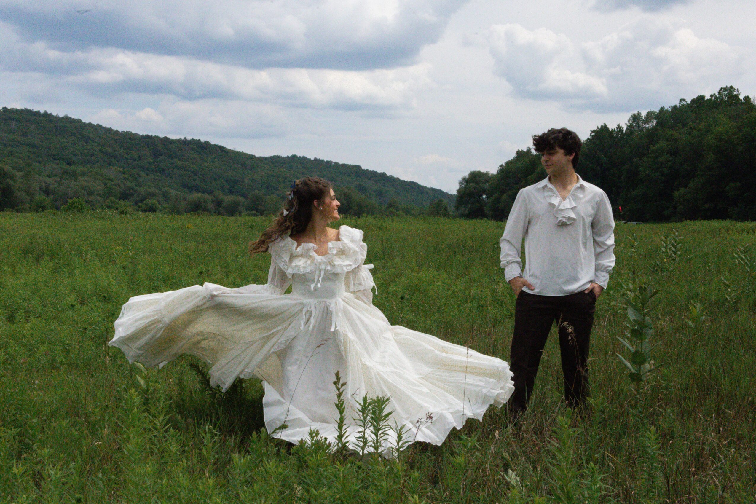 A woman in a white dress twirls in a grassy field with a man in a white shirt nearby, mountains and clouds in background.