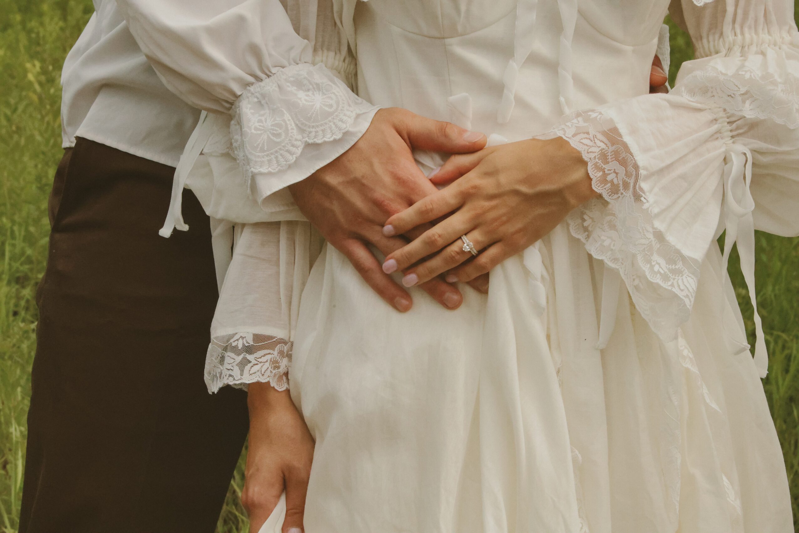 Two people holding hands, one wearing a white dress with lace details, the other in dark pants, outdoors.