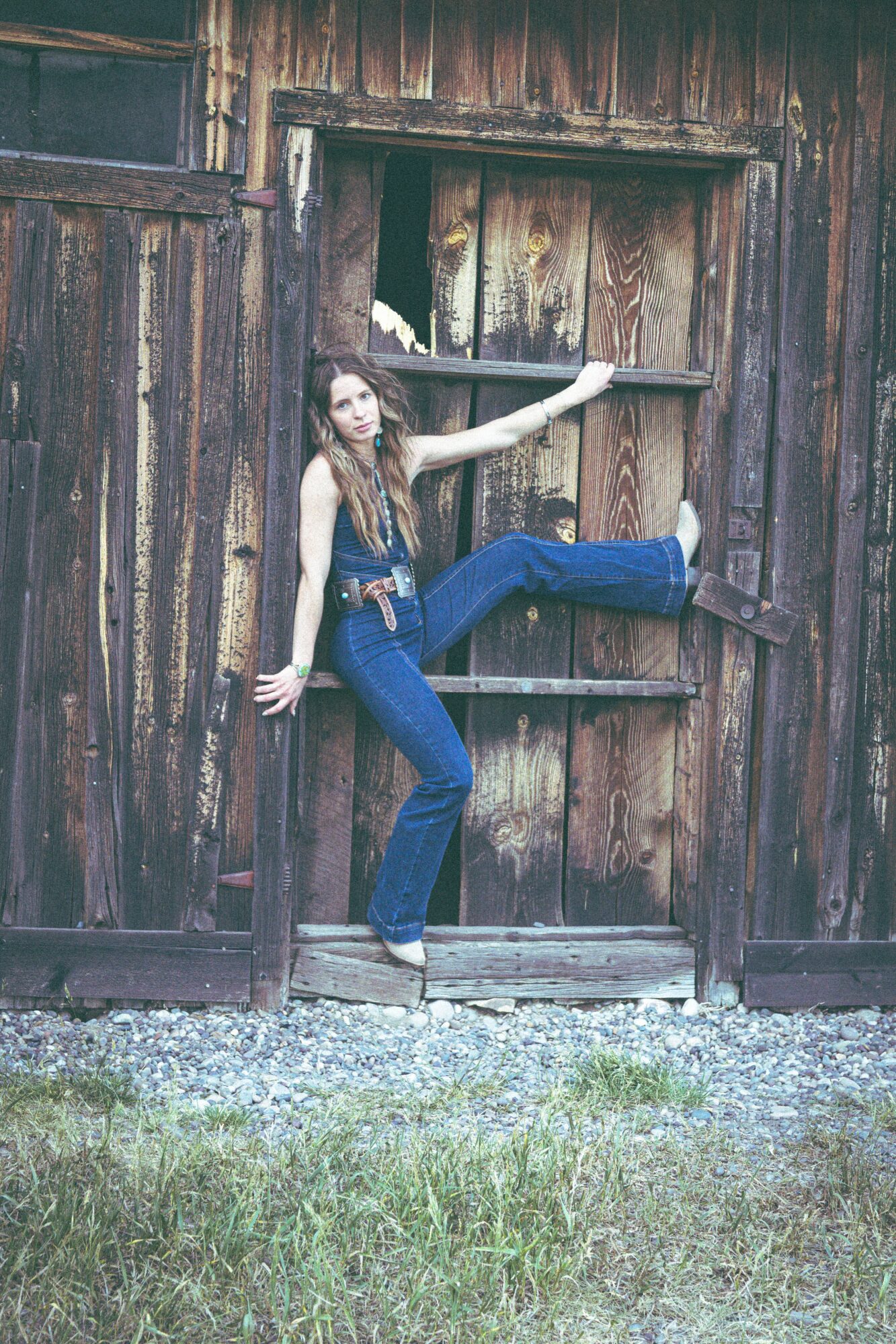 Young woman in jeans and a black top poses with one leg extended through a window of a weathered wooden building.