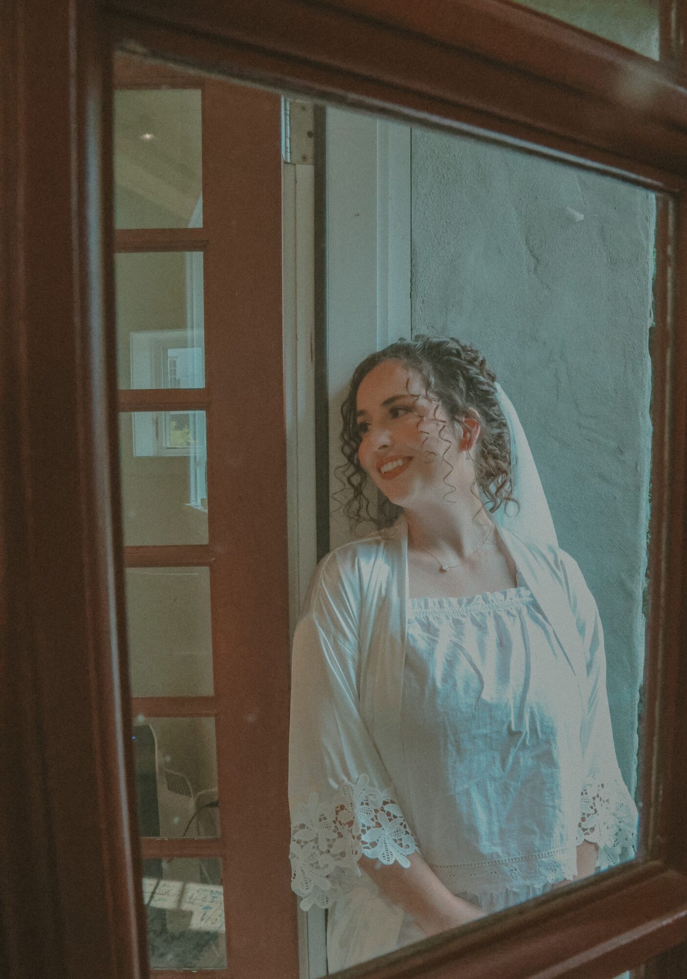 Young woman with curly hair wearing a white dress, smiling and looking to the side, standing near a window.