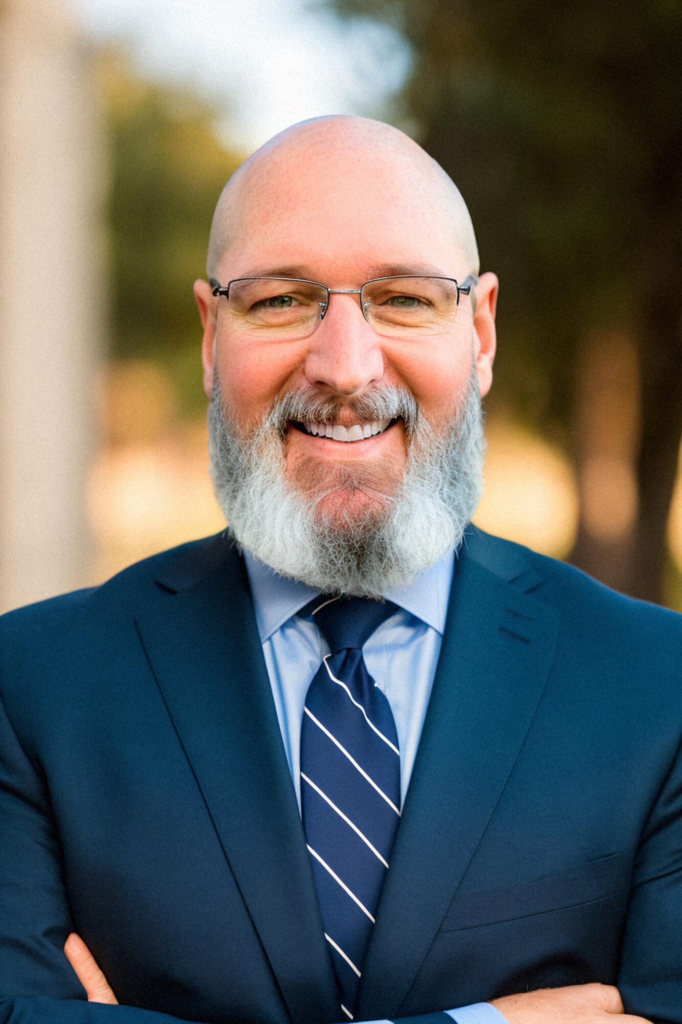 Smiling man with glasses, bald head, and beard, wearing a suit and tie outdoors.
