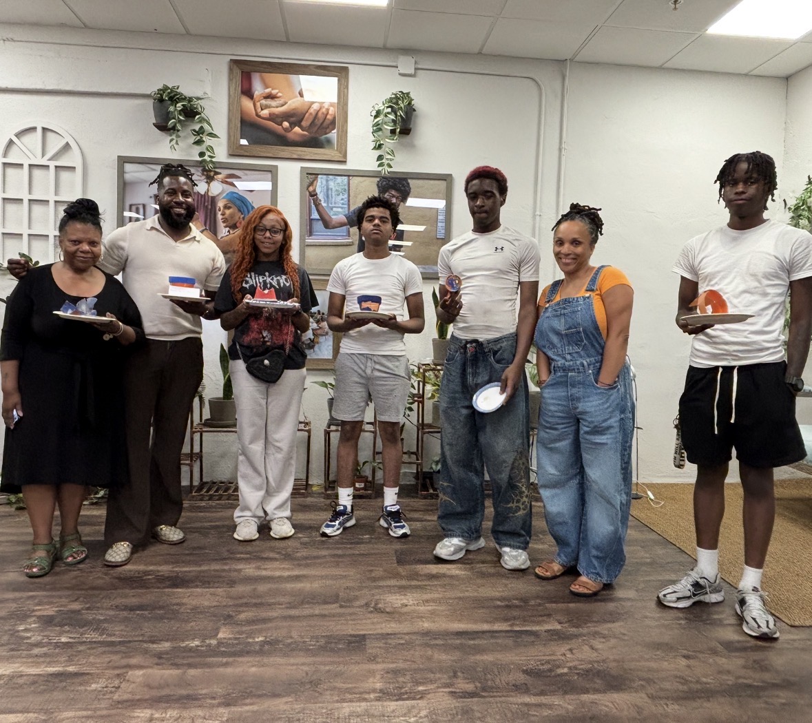 Group of eight people standing indoors, some holding plates, in front of a decorated wall with framed pictures.