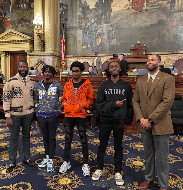 Five people standing in a room with ornate decor, paintings, and a large mural, posing for a photo.