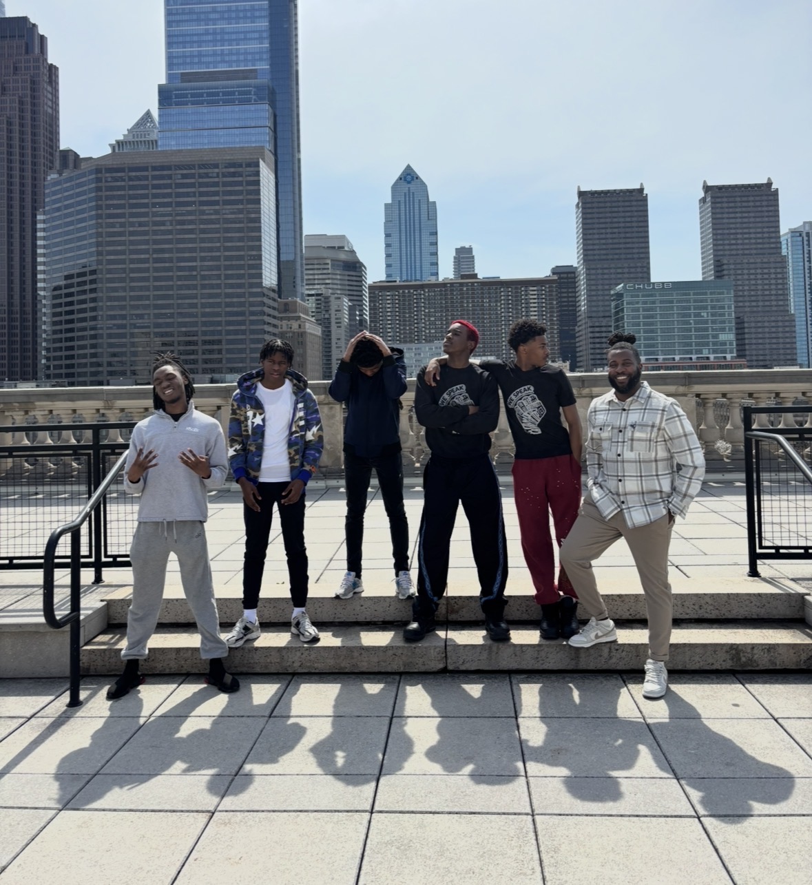 Six people standing on a city rooftop with skyscrapers in the background, some smiling and talking, shadows cast on the ground.