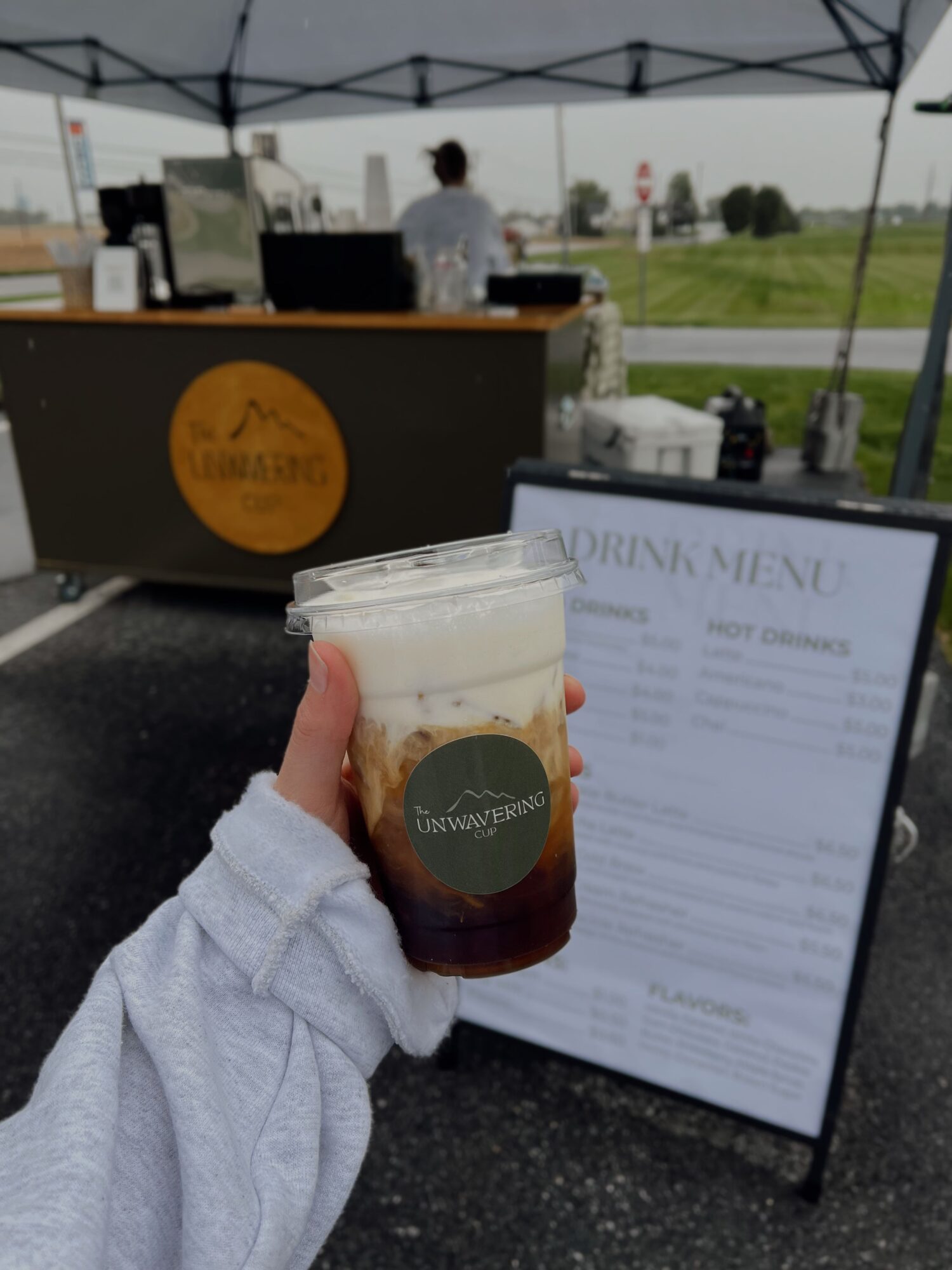 Person holding iced coffee with a straw, outdoor setting with menu board and people in background.