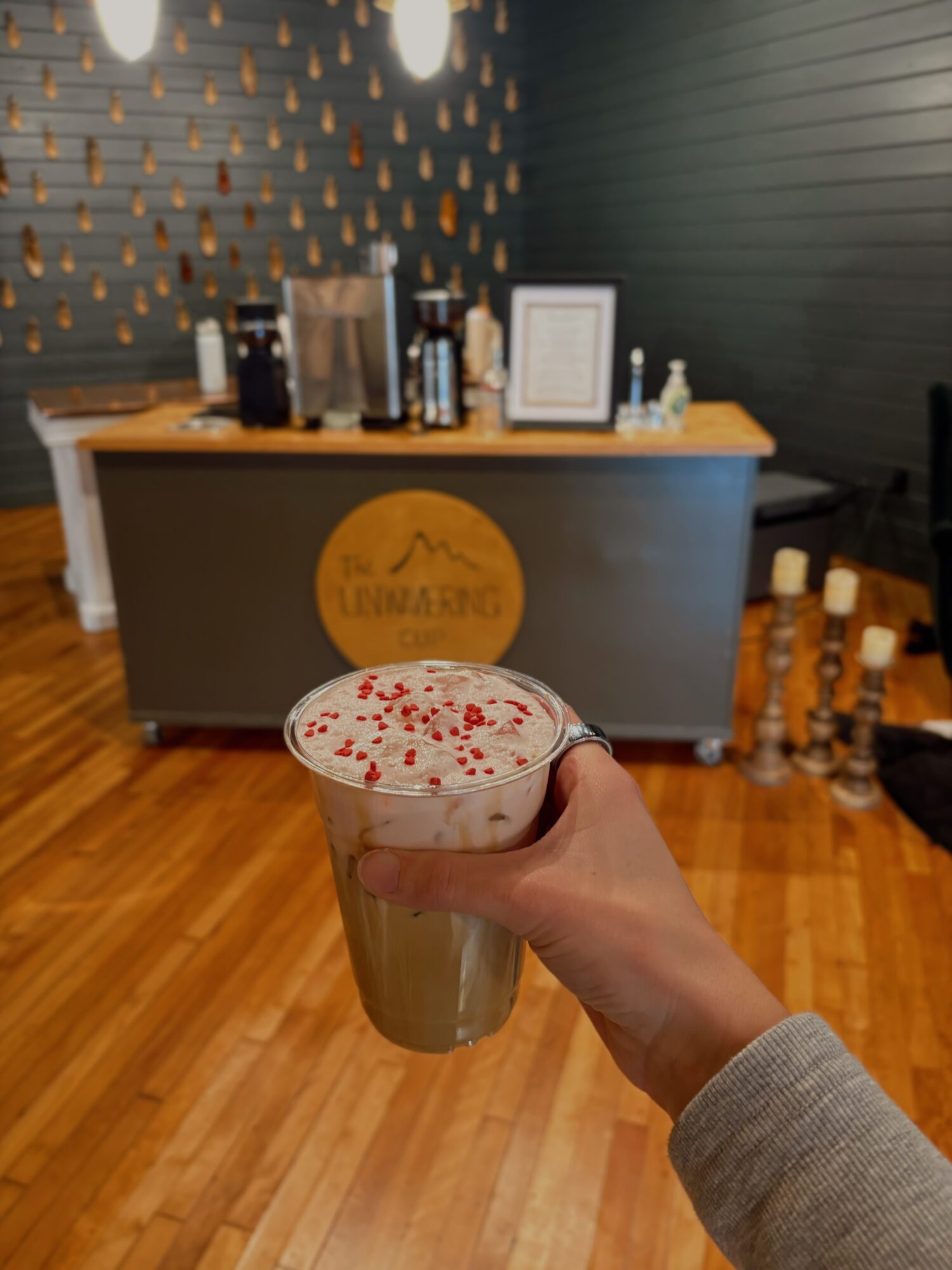 Person holding a drink with foam and red sprinkles in front of a coffee stand, wooden floor, and dark wall.