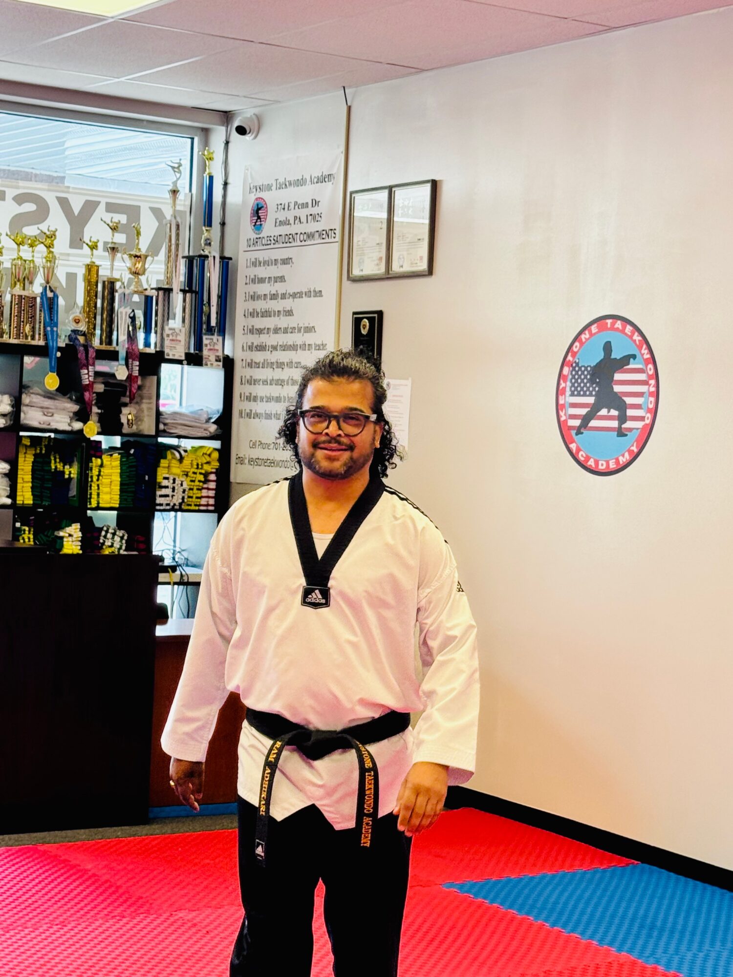Person in taekwondo uniform standing on a red and blue mat, smiling indoors.