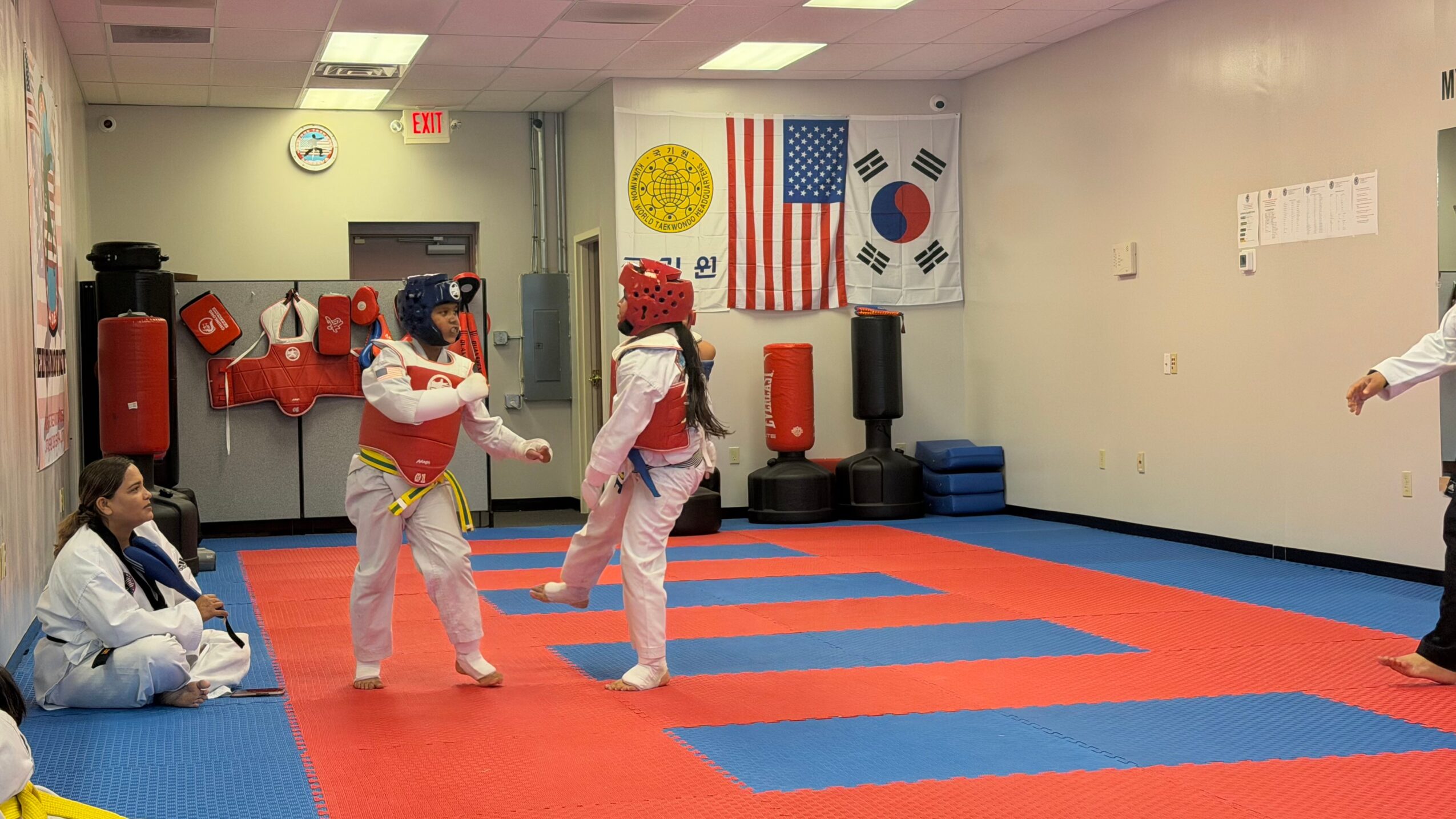 Two children in taekwondo uniforms spar in a martial arts dojo with flags on the wall, a punching bag, and mats.