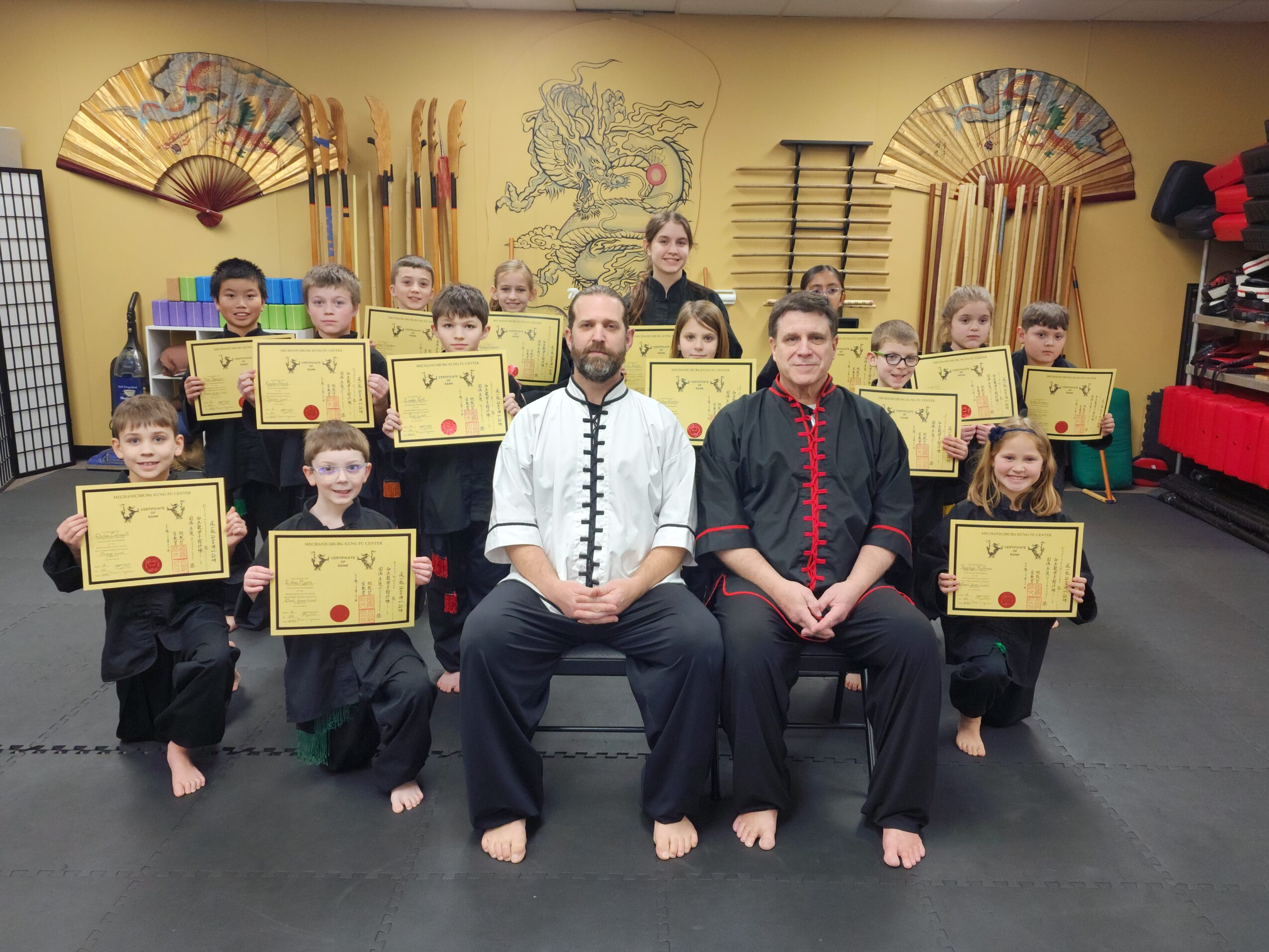 Group of children and two instructors in martial arts uniforms holding certificates, in a dojo with decorative fans and weapons on the wall.