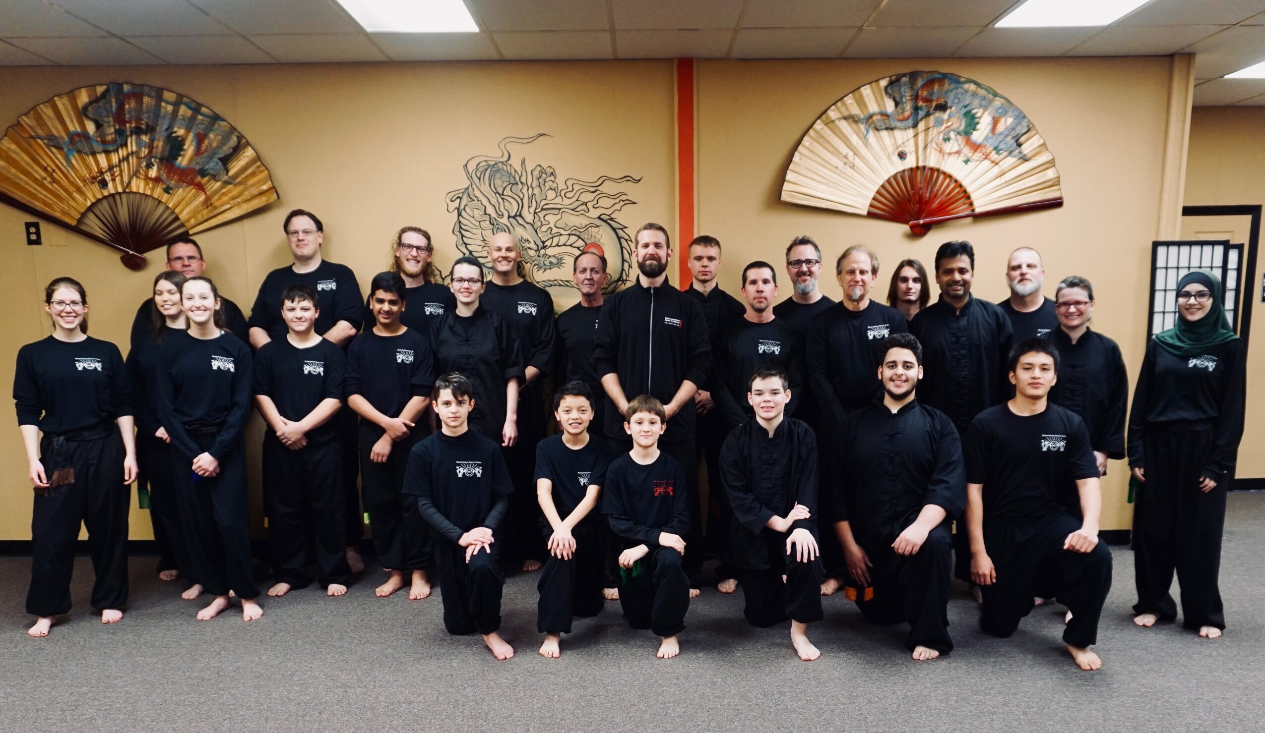 Group of people in black uniforms posing indoors with decorative fans on the wall behind them.