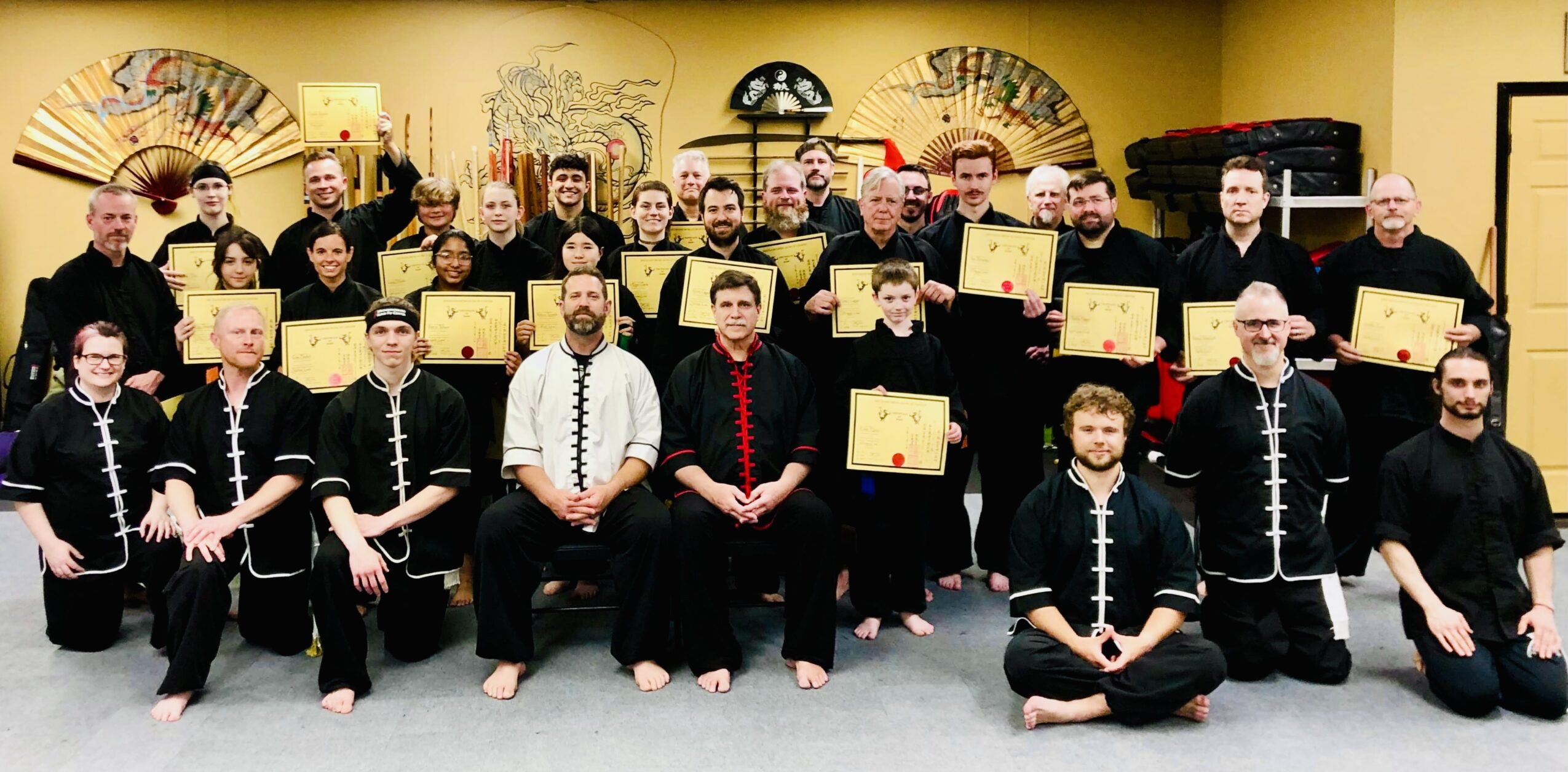 Group of people in traditional attire holding certificates, standing in front of a yellow wall with decorative fans and artwork.