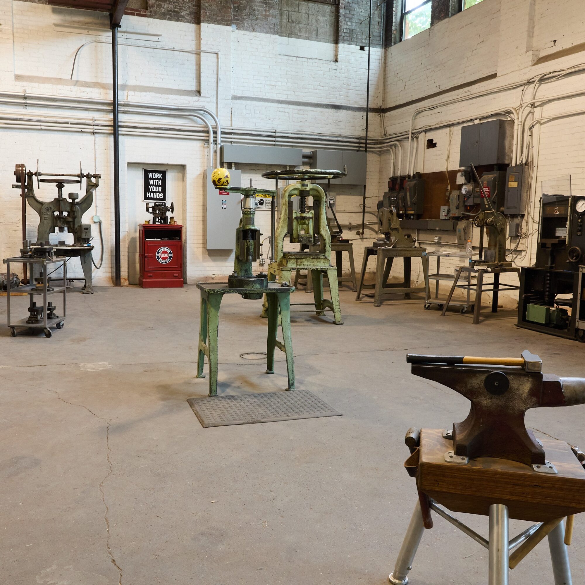 Workshop with various metalworking tools and equipment, including a workbench, machines, and an anvil, in a spacious industrial setting.