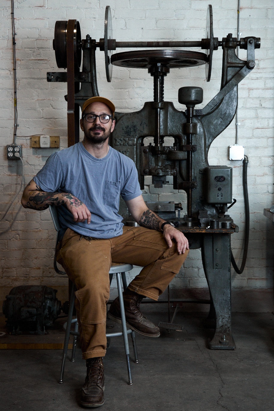 Man with glasses and tattoos sitting on a stool in front of large industrial machine against a white brick wall.