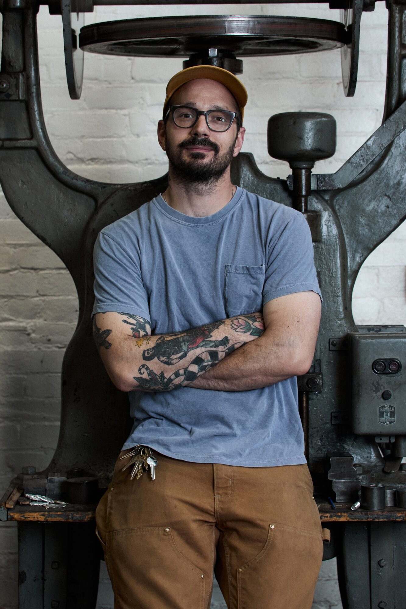 Man with glasses, beard, tattoos, wearing a yellow cap and blue t-shirt, standing with arms crossed in front of industrial machinery.
