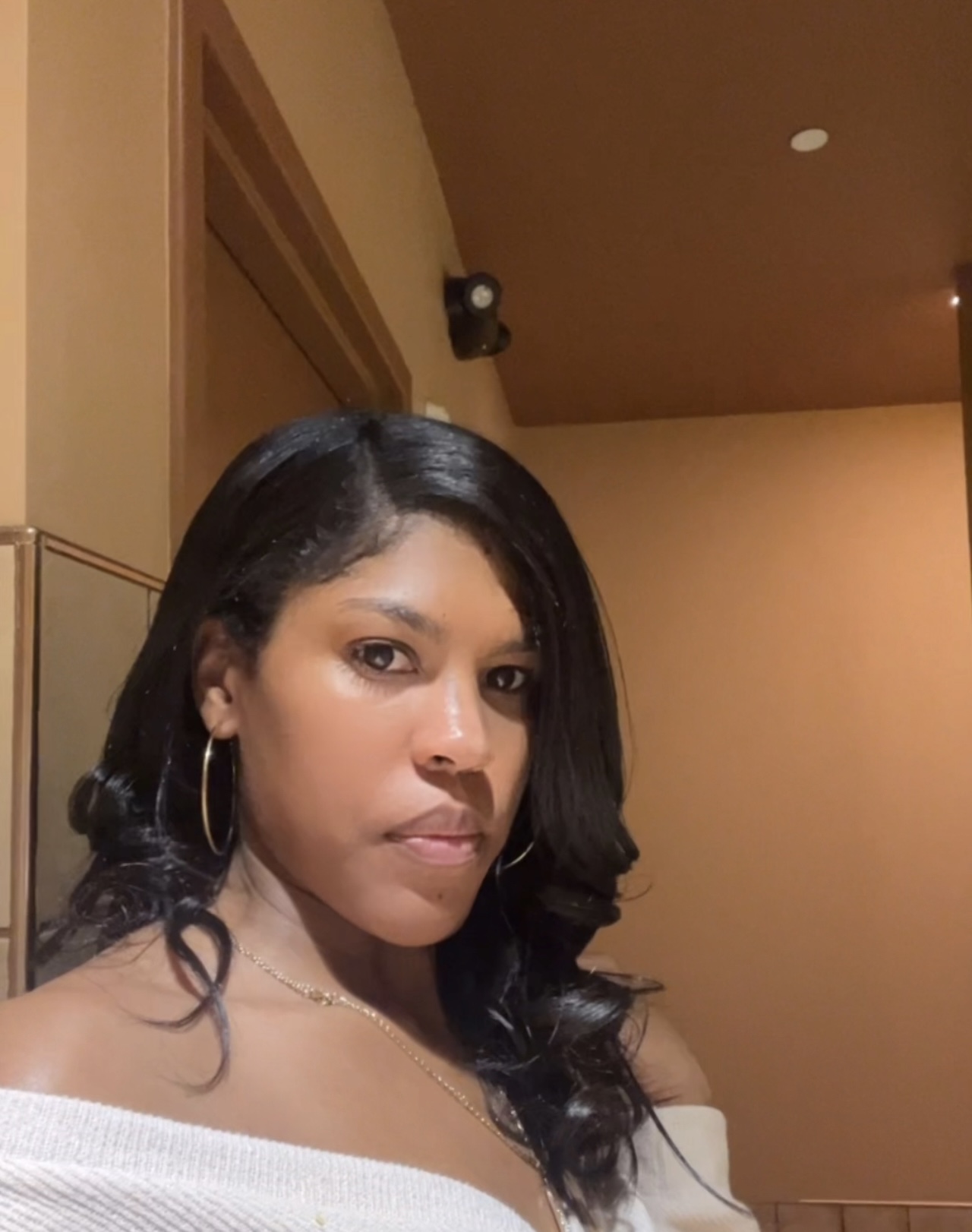 Woman with long black hair, wearing hoop earrings and a white off-shoulder top, looking at the camera indoors.