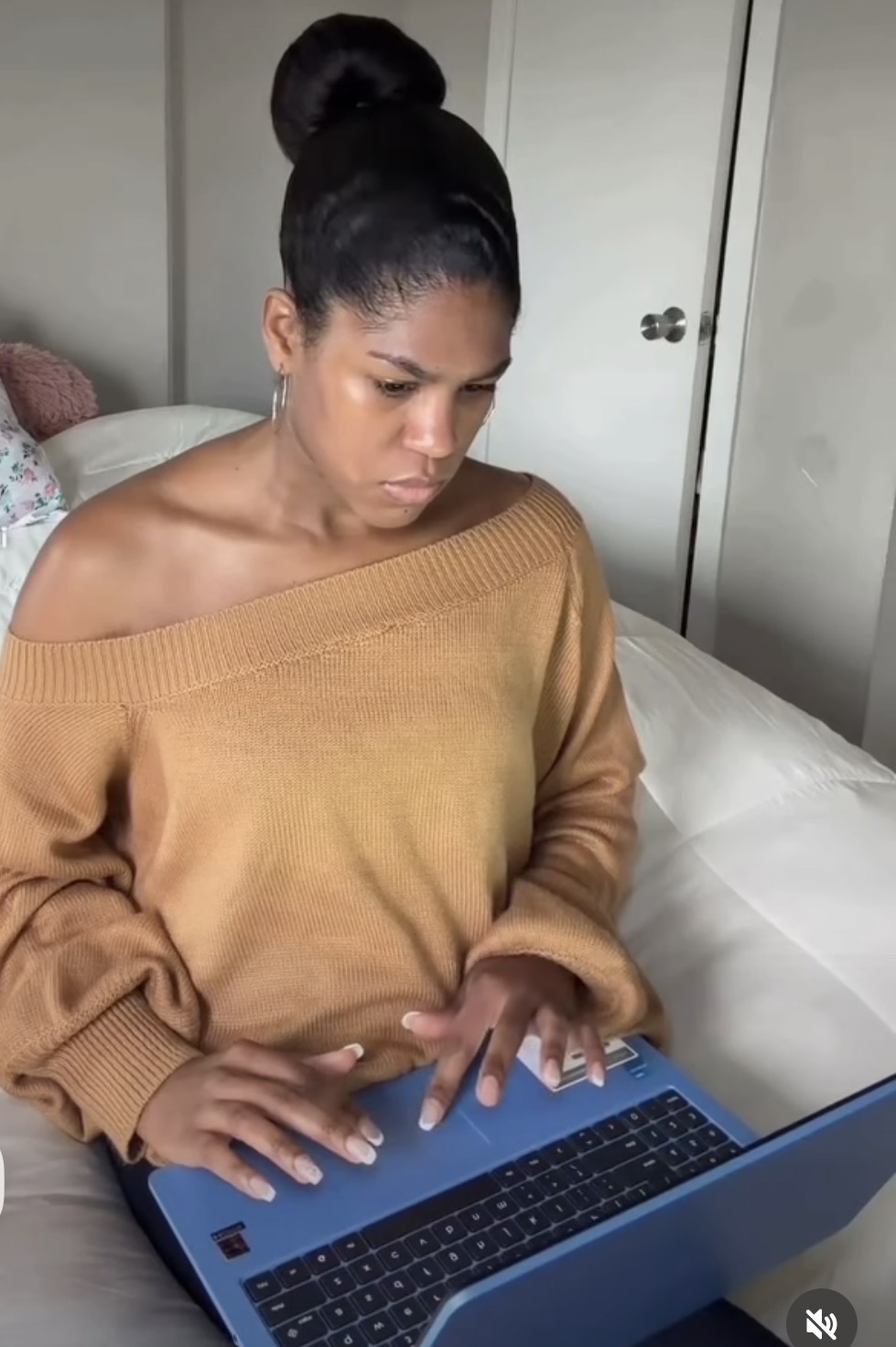 Woman with dark hair in a bun, wearing a brown off-shoulder sweater, sitting on a bed using a blue laptop.