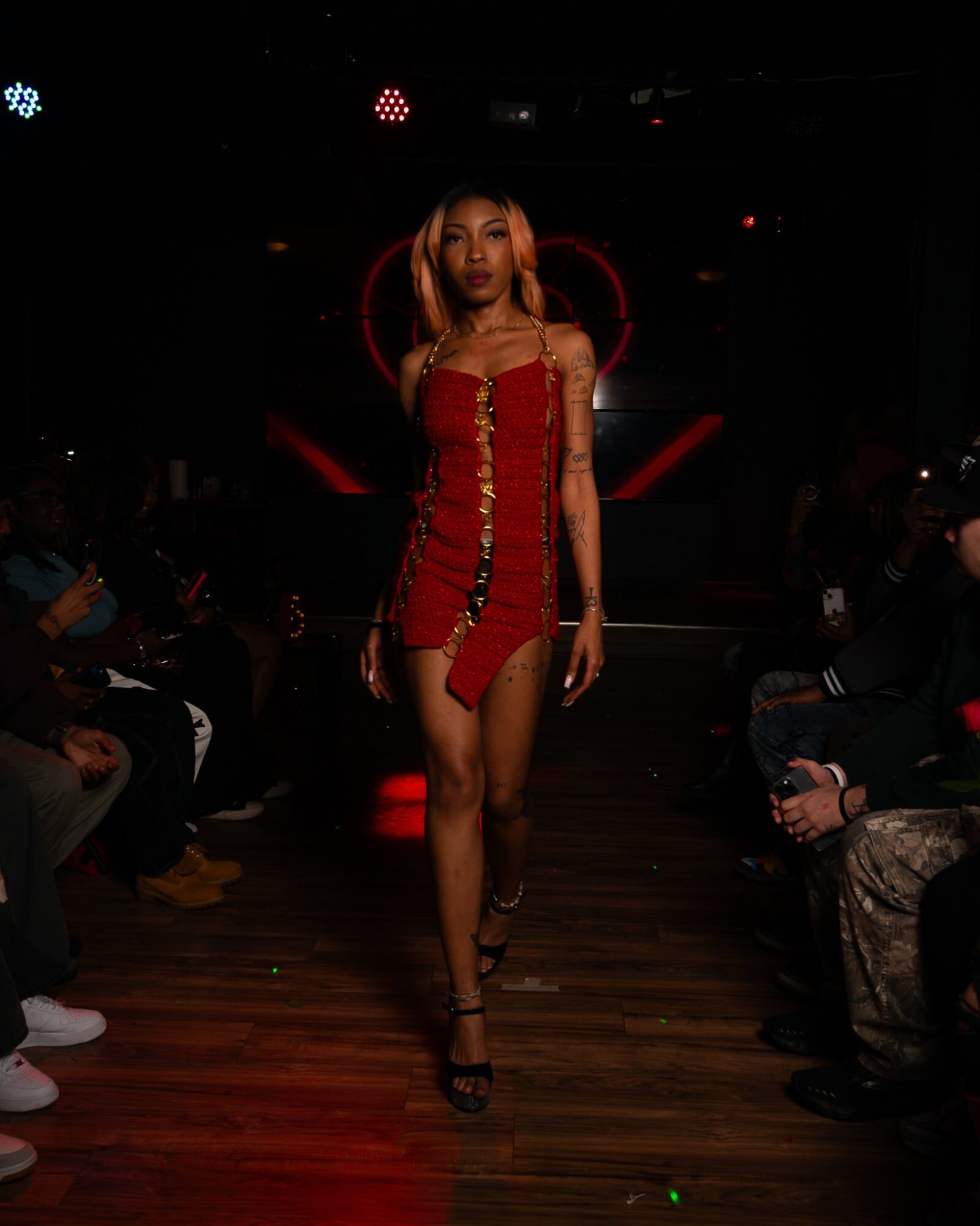 A woman walks on a runway in a red dress with gold buttons, surrounded by seated audience in a dark setting.