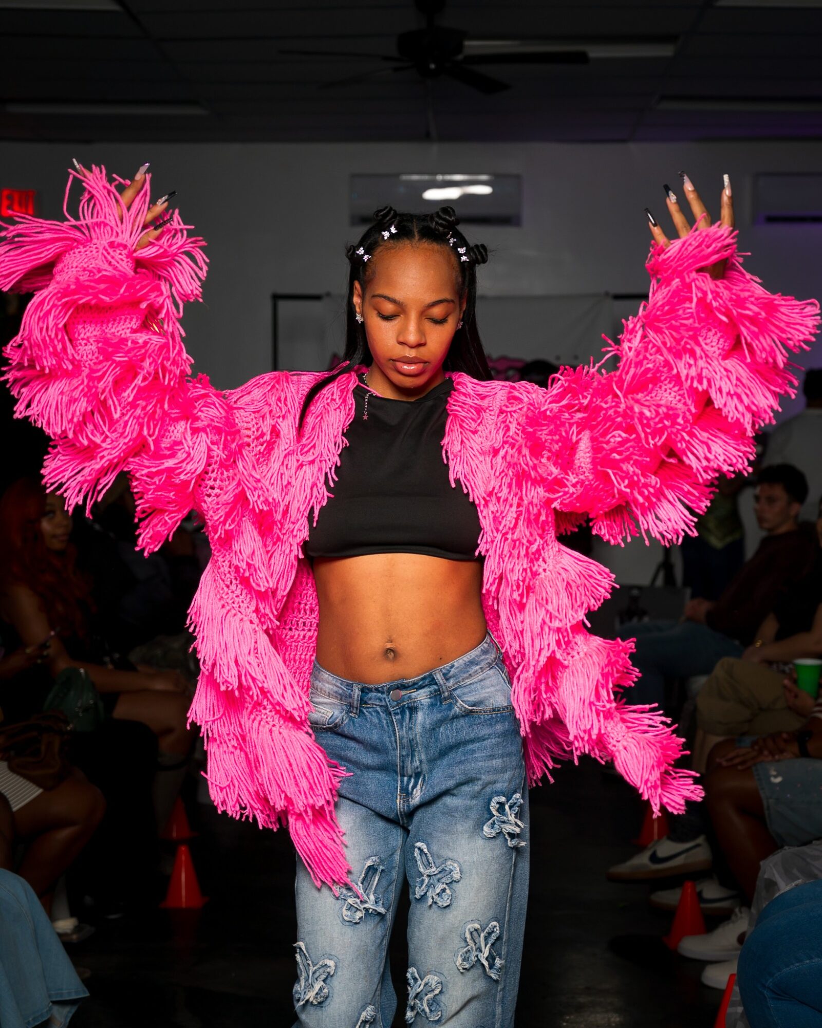 Young girl with arms raised, wearing a bright pink fluffy jacket, black crop top, and ripped jeans, in a room with people.