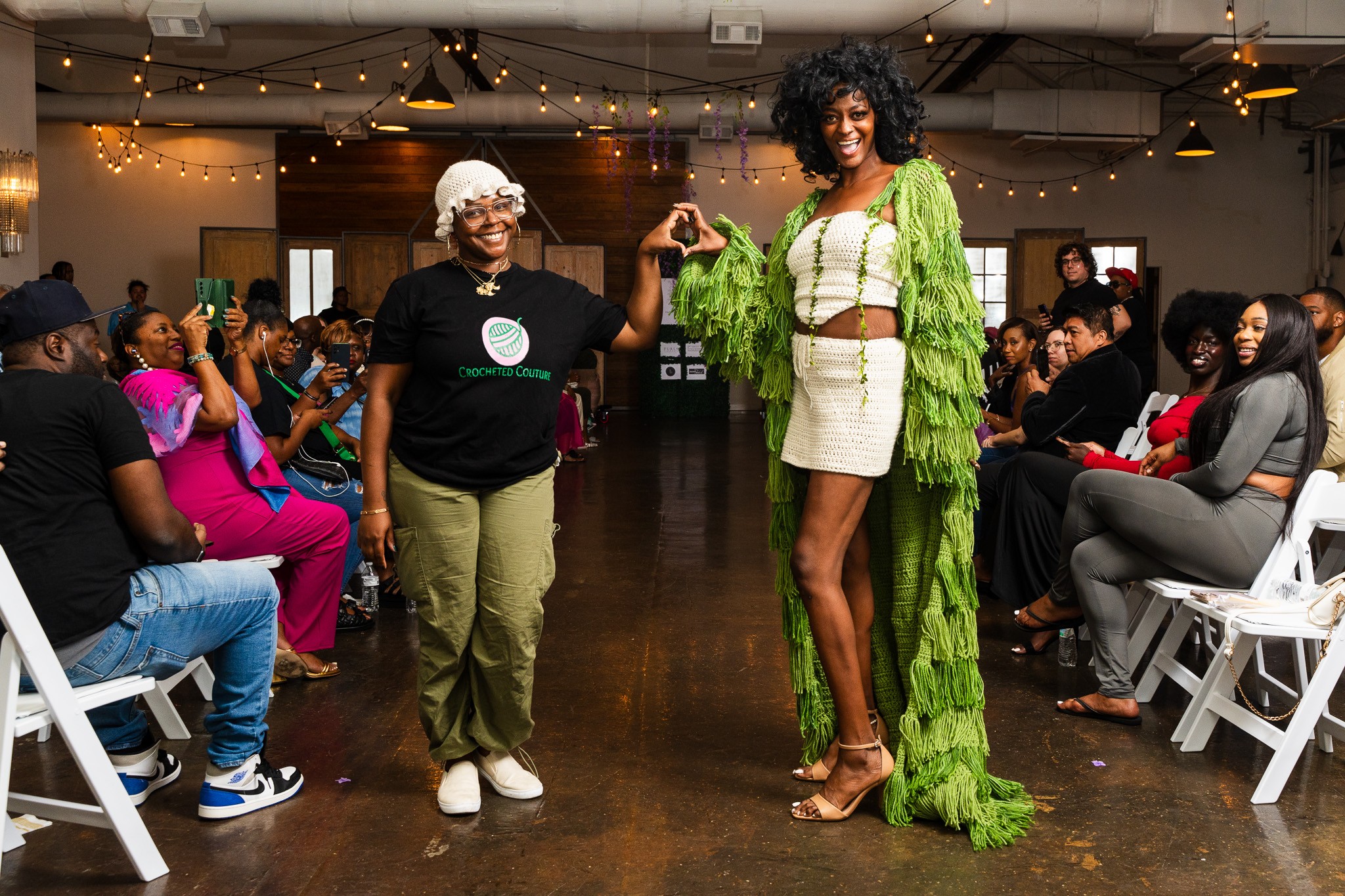 Two women stand in the center of a room with seated audience on both sides, one taking a photo of the other.