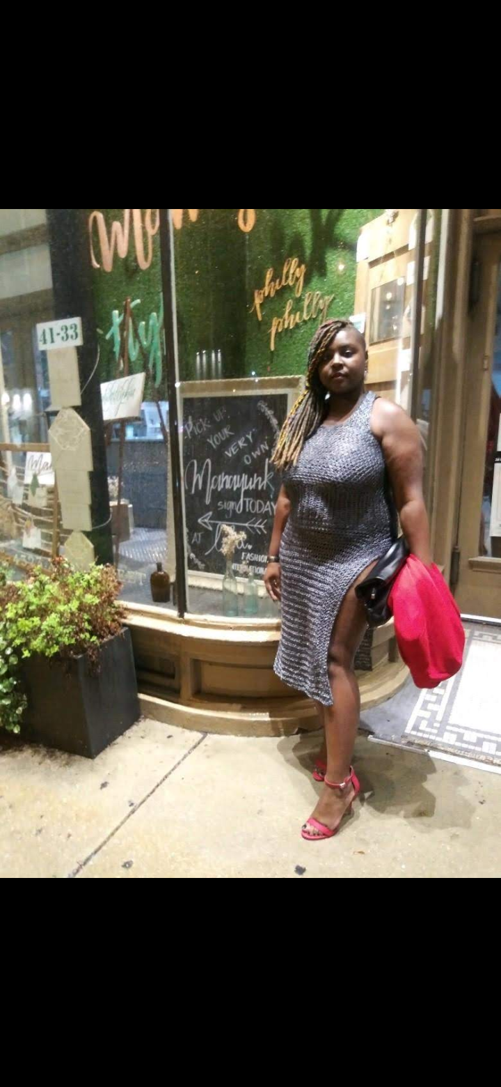 Woman standing outside a shop holding a red bag, wearing a dress and sandals, with a chalkboard sign behind her.