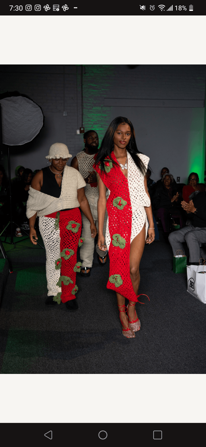 Group of people walking on runway during fashion show, woman in red dress with floral scarf, others in stylish outfits, audience seated