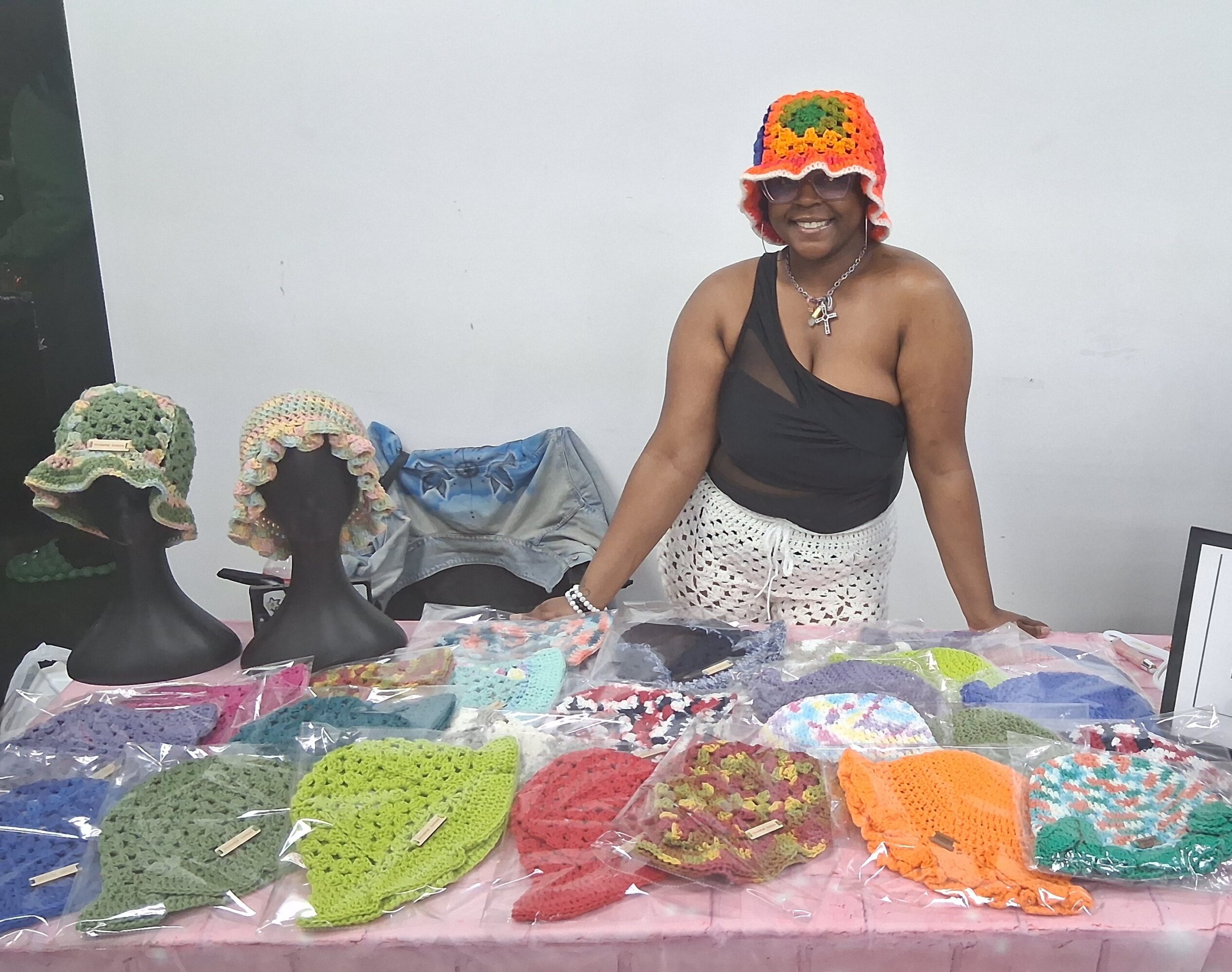 Woman wearing a colorful hat, black top, and white skirt, standing behind a table with handmade crochet items.