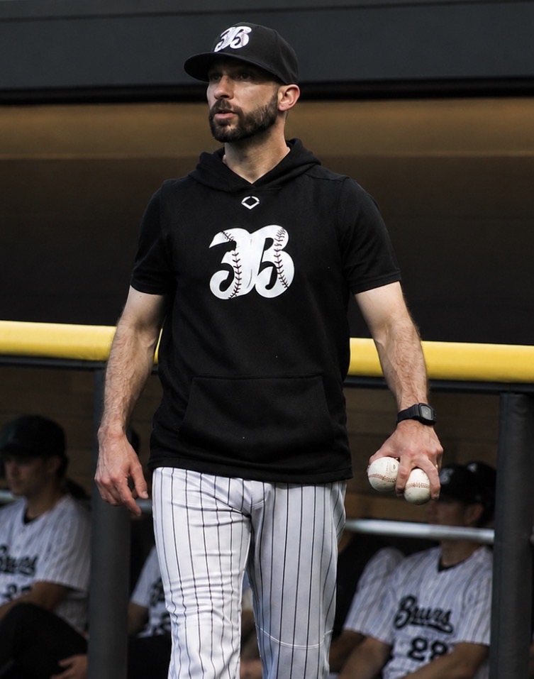 Man in baseball uniform holding baseballs, wearing a black hoodie, cap, and striped pants, standing near a yellow railing.