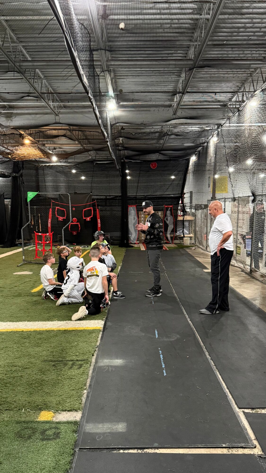 Indoor sports facility with children sitting on the floor and adults standing nearby, artificial turf and netting visible.