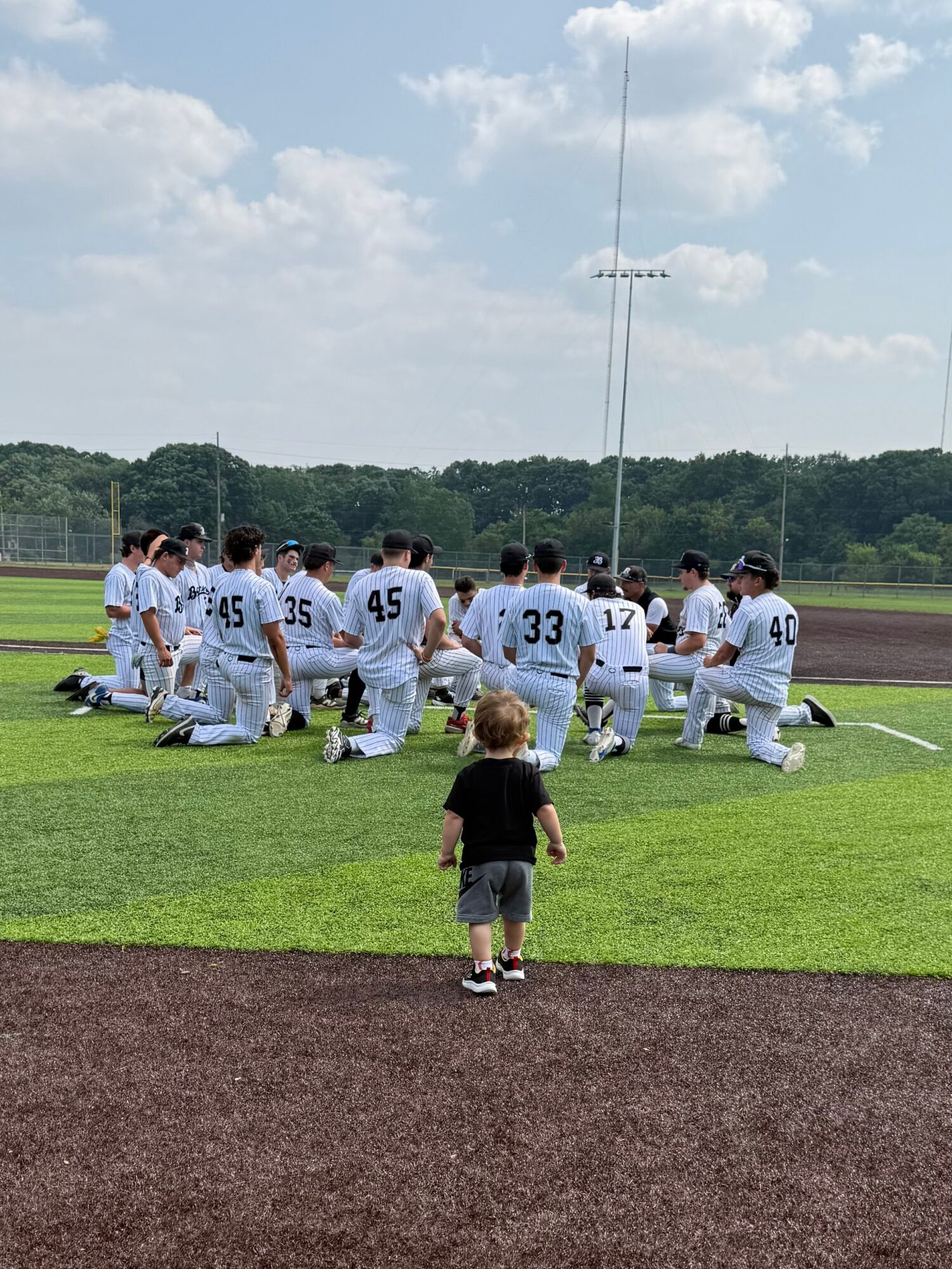 Group of baseball players kneeling on the field, with a child standing in front, on a sunny day.