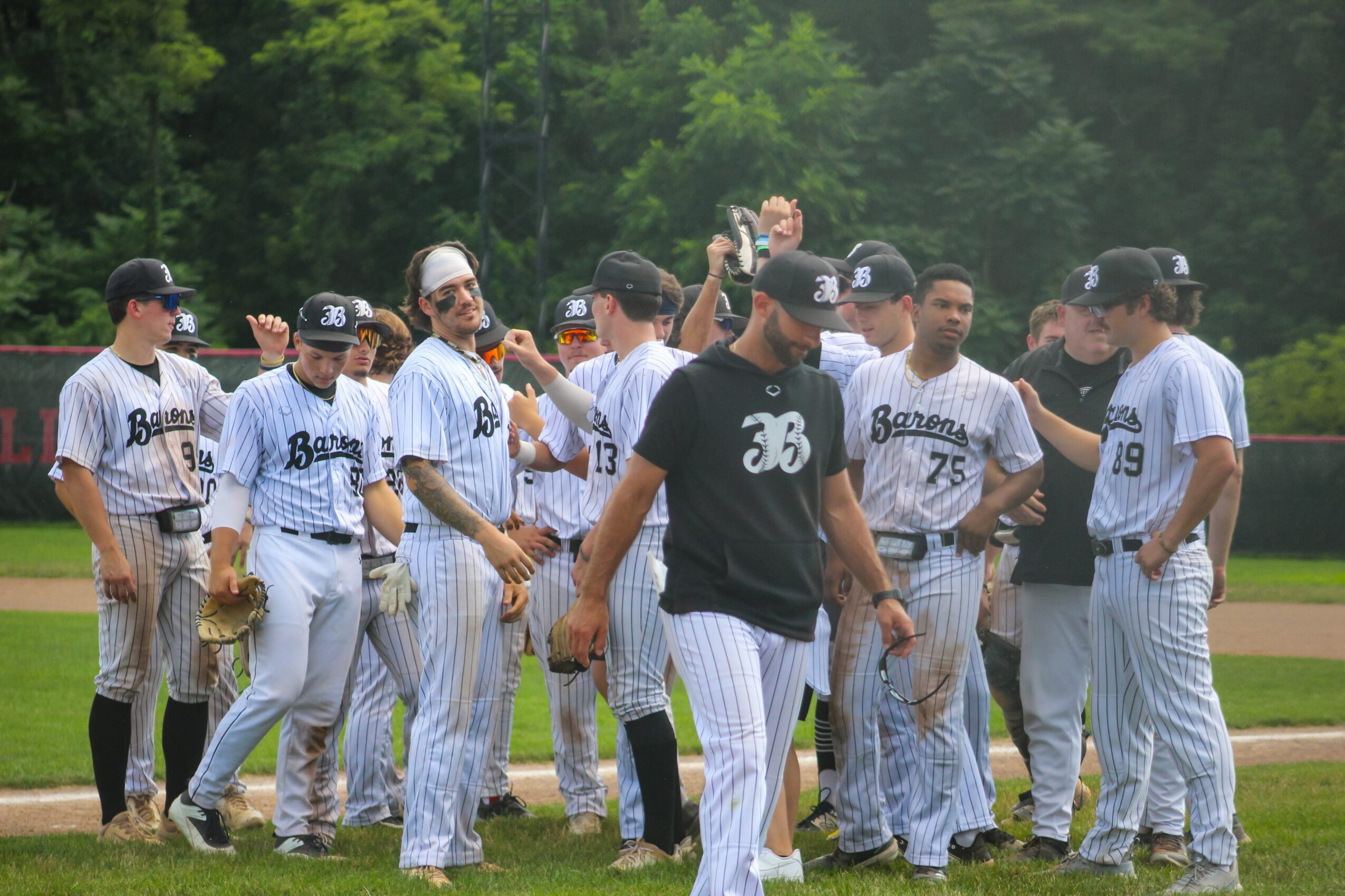 Group of baseball players in uniform standing on a field, some high-fiving and talking, with trees in the background.