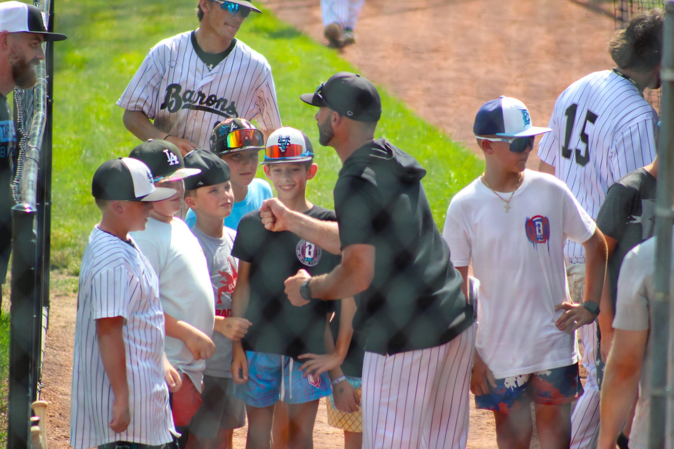 Group of children and adults gathered outdoors, some wearing baseball jerseys and caps, with a coach or adult giving instructions.