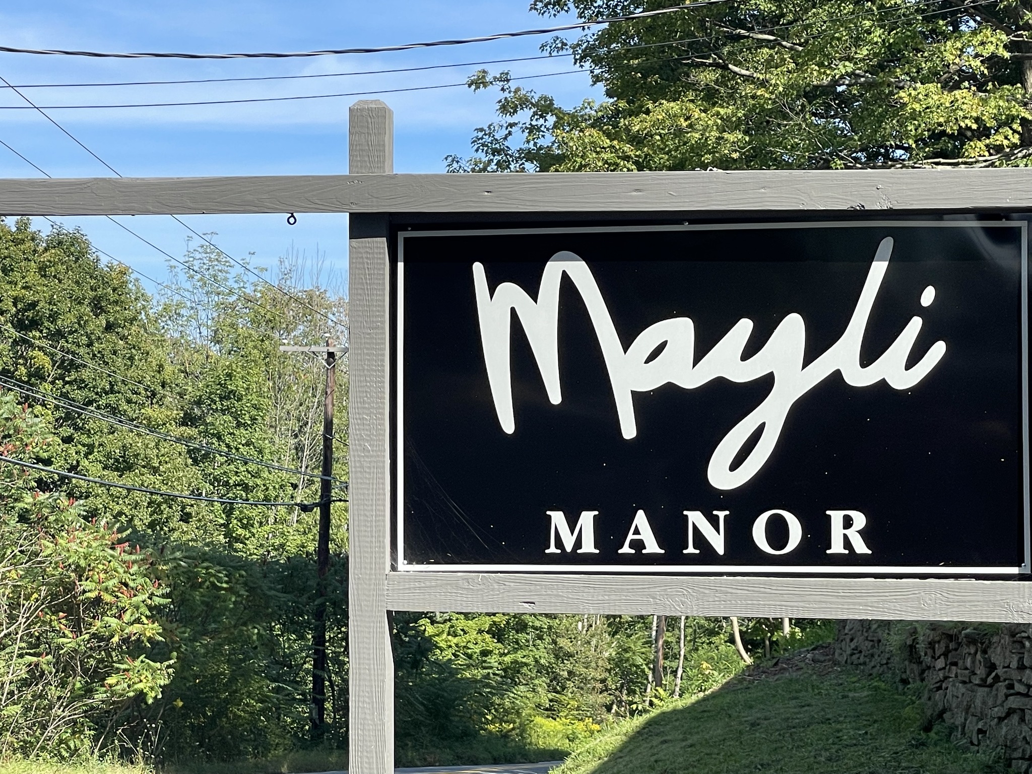 Sign reading 'Mayli MANOR' with trees and blue sky in background.