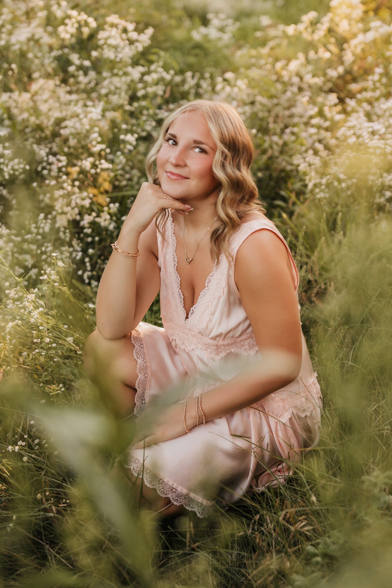 Young woman with wavy blonde hair sitting outdoors among greenery and white flowers, smiling gently, wearing a light pink dress.
