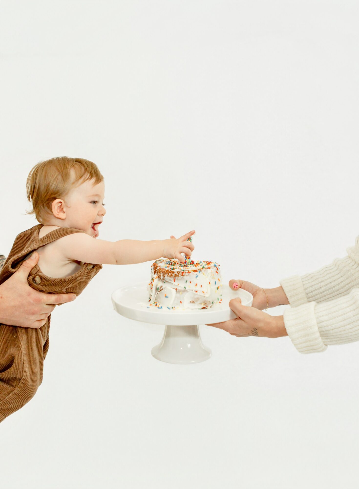 Child reaching for a cake on a stand held by an adult's hands, with a plain white background.