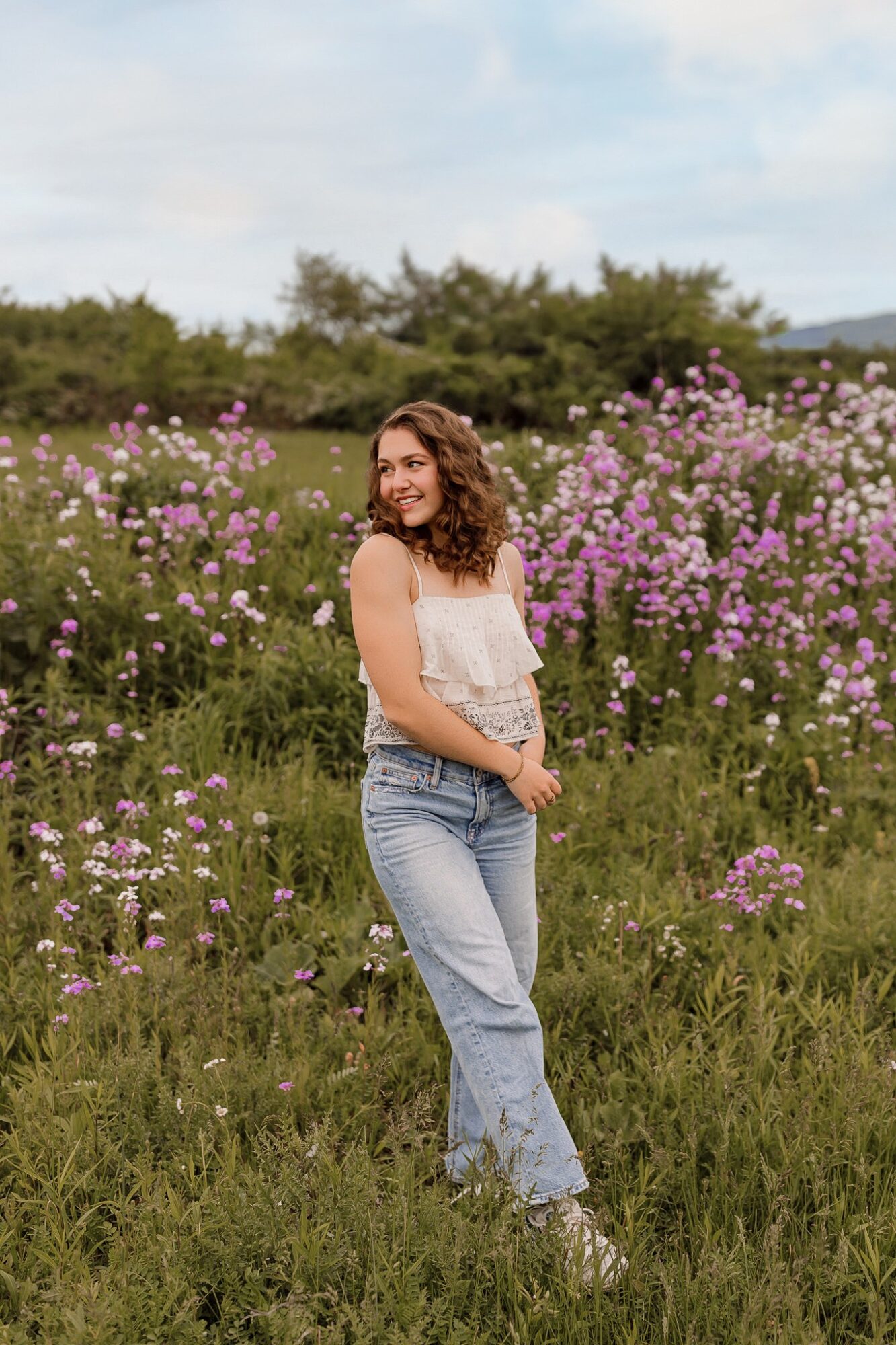 Young woman with curly hair smiling in a field of pink and purple flowers, wearing a white top and jeans.