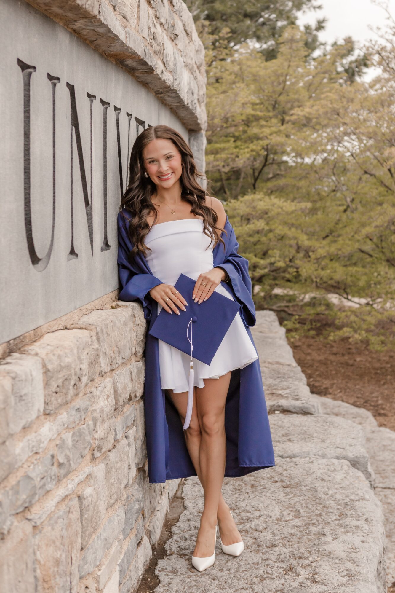 Young woman in graduation gown and cap, smiling, standing outdoors near a stone wall with trees in background.