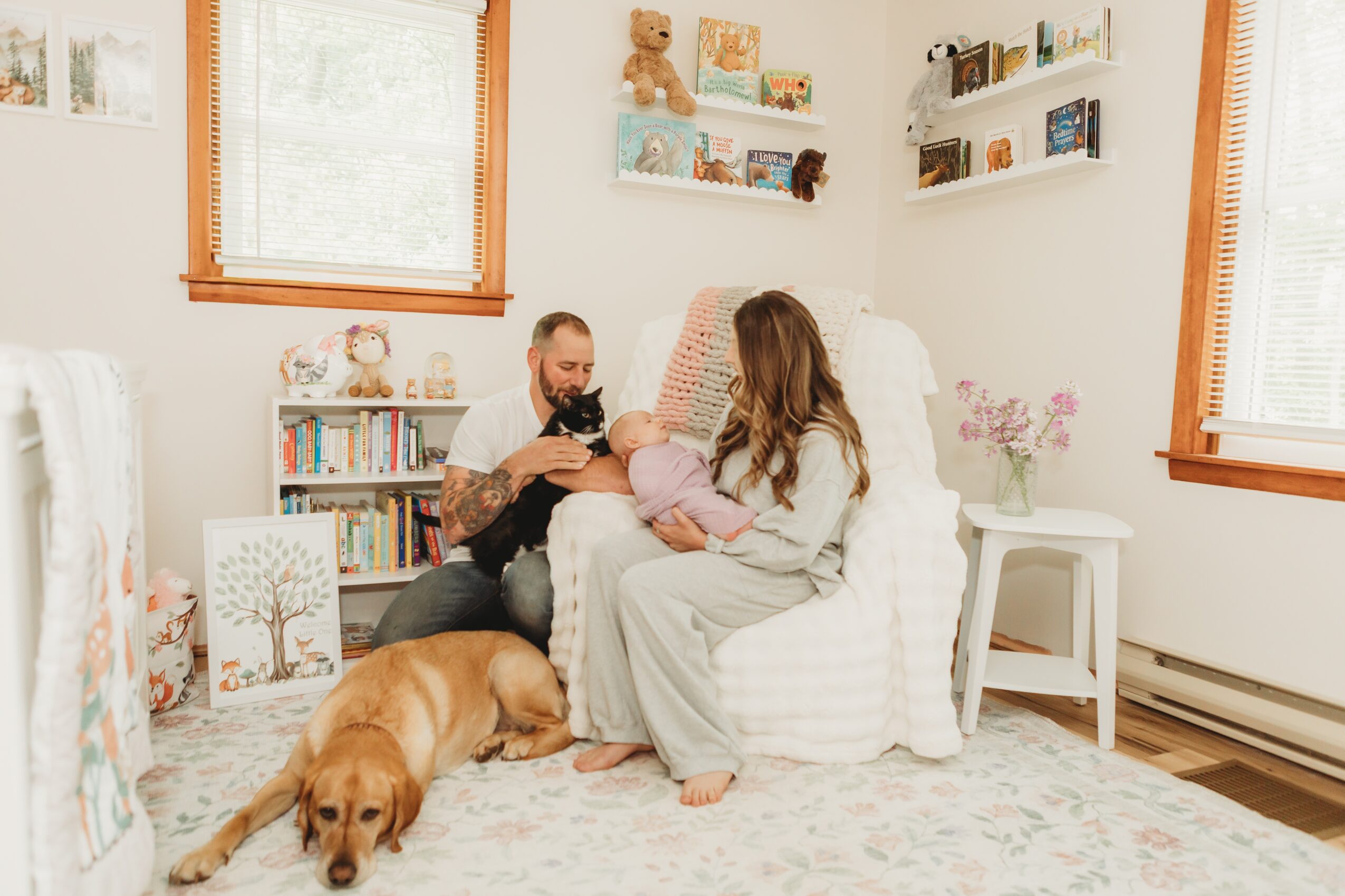 Family with a woman, man, baby, and dog in a cozy living room with bookshelves and windows, sitting on a white armchair.