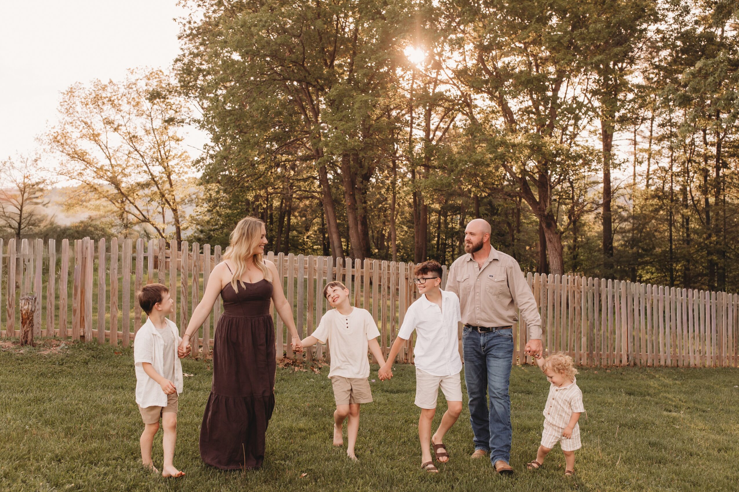 Family of six holding hands outdoors on grass with trees and a fence in the background, sunset lighting.