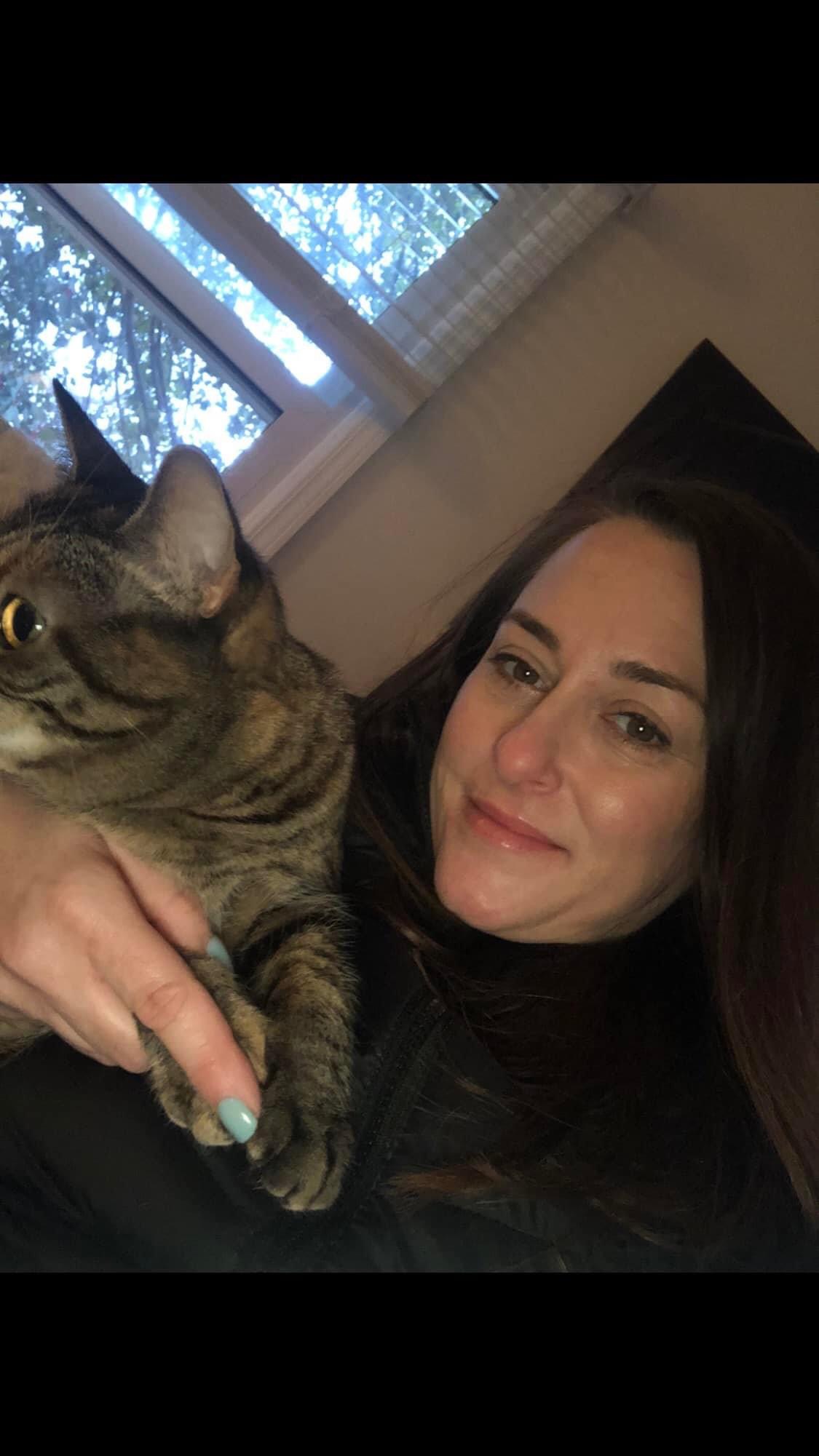 Woman with dark hair smiling, holding a tabby cat near a window with trees outside.