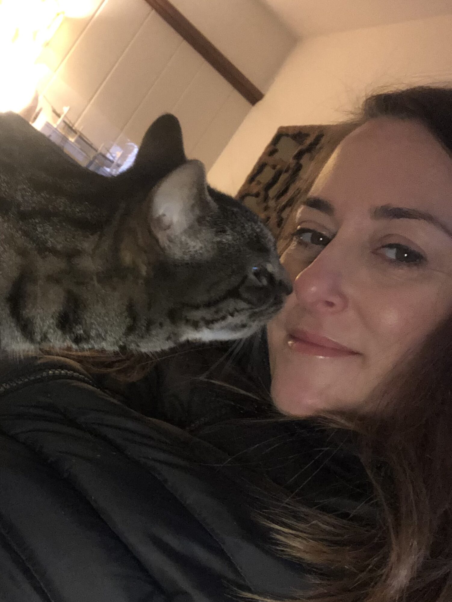 Person smiling with a cat close to their face indoors, warm lighting, cozy setting.