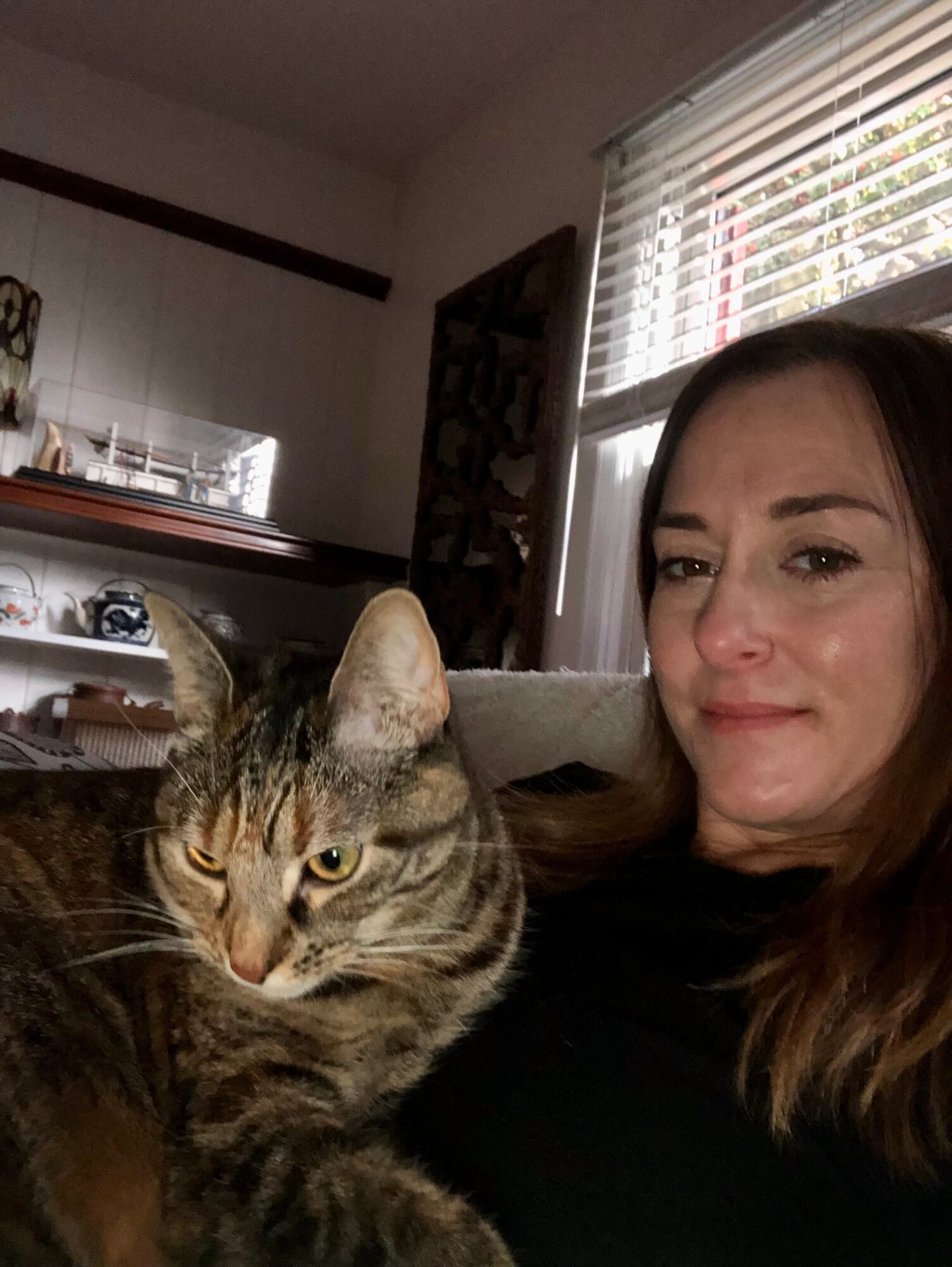 Woman with long brown hair sitting indoors next to a tabby cat near a window with blinds.
