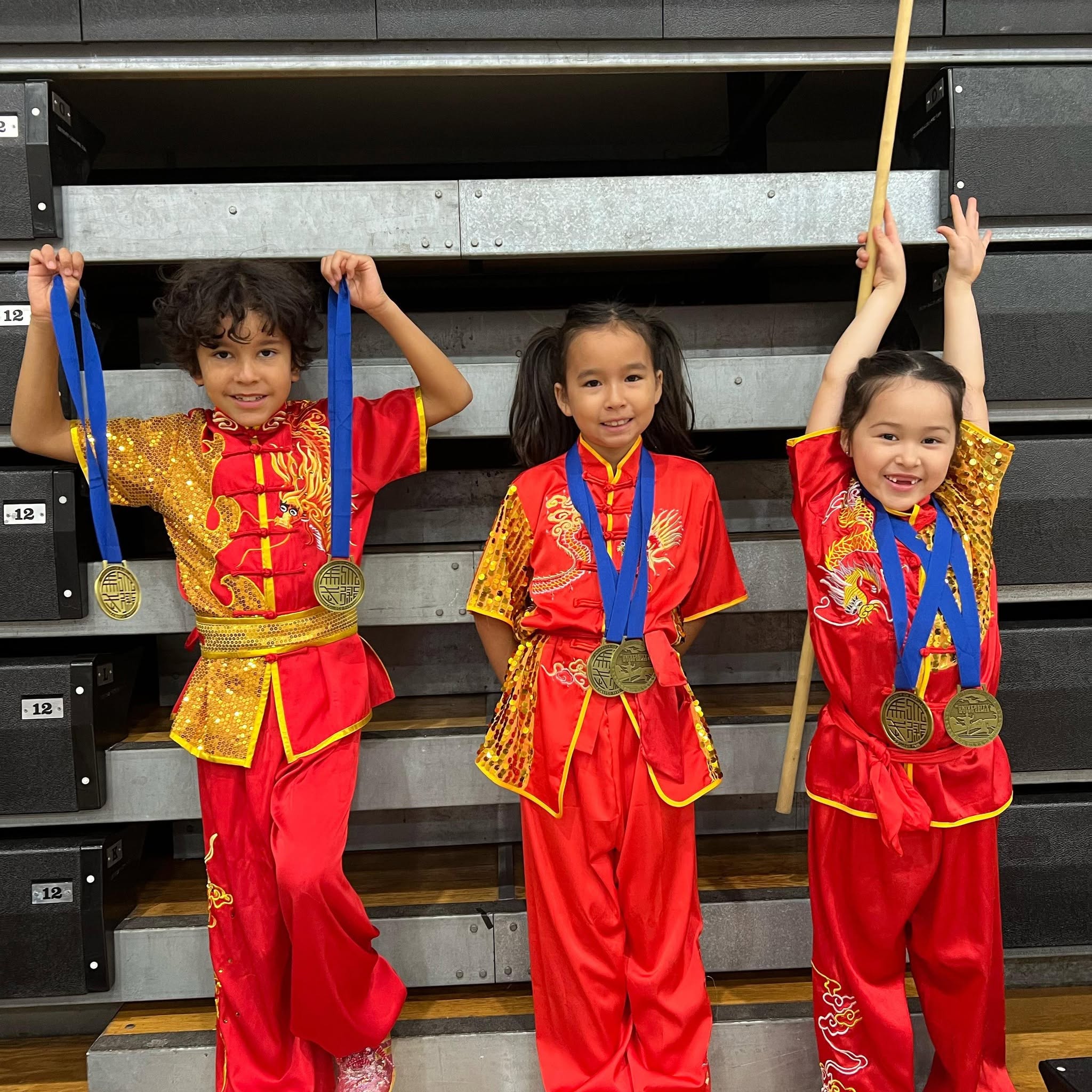 Three children in red and yellow martial arts uniforms holding medals and a staff, smiling, standing in front of a black and gray background.