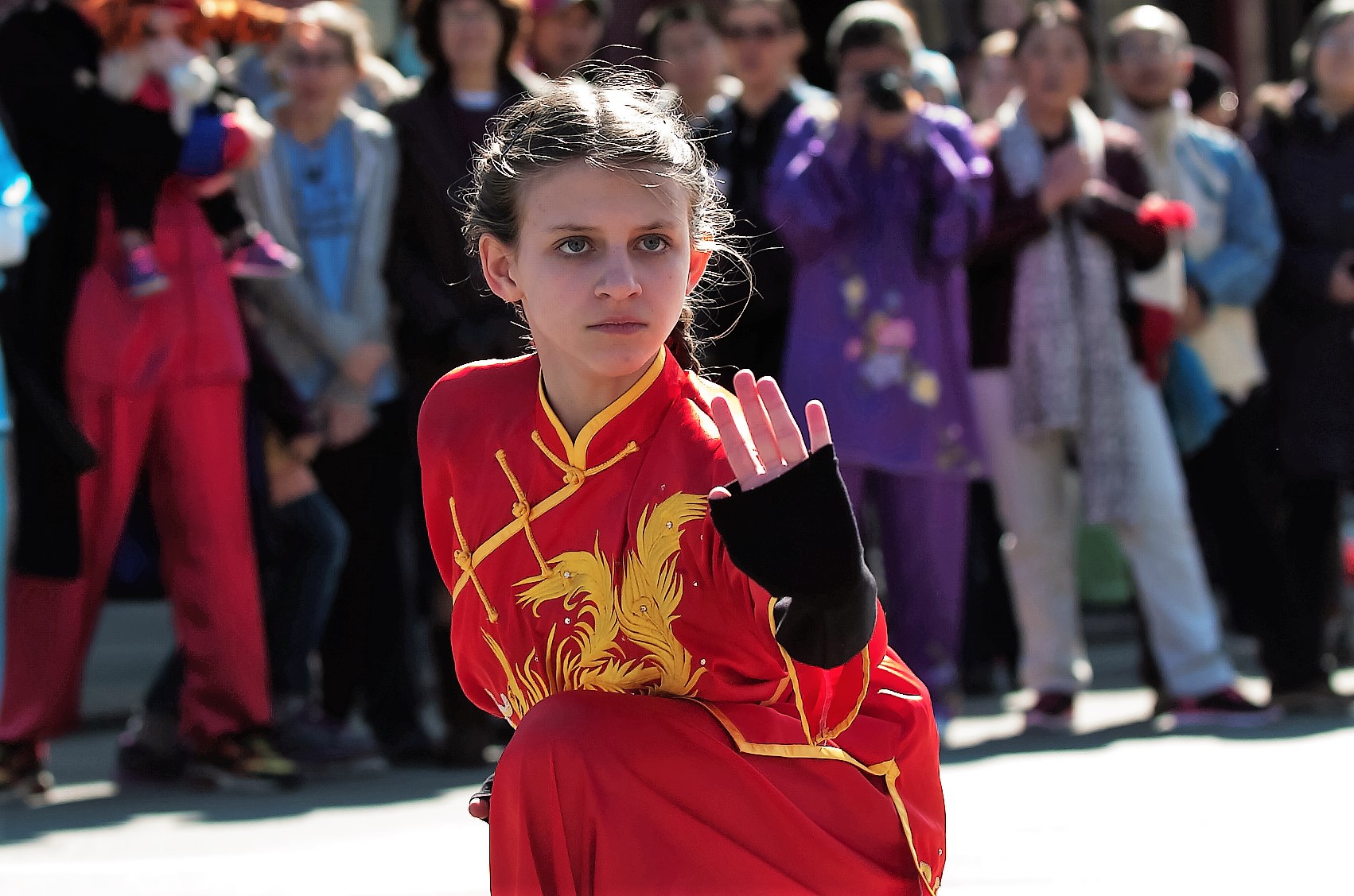 Young girl in red martial arts uniform performing a move outdoors, with a crowd watching behind her.