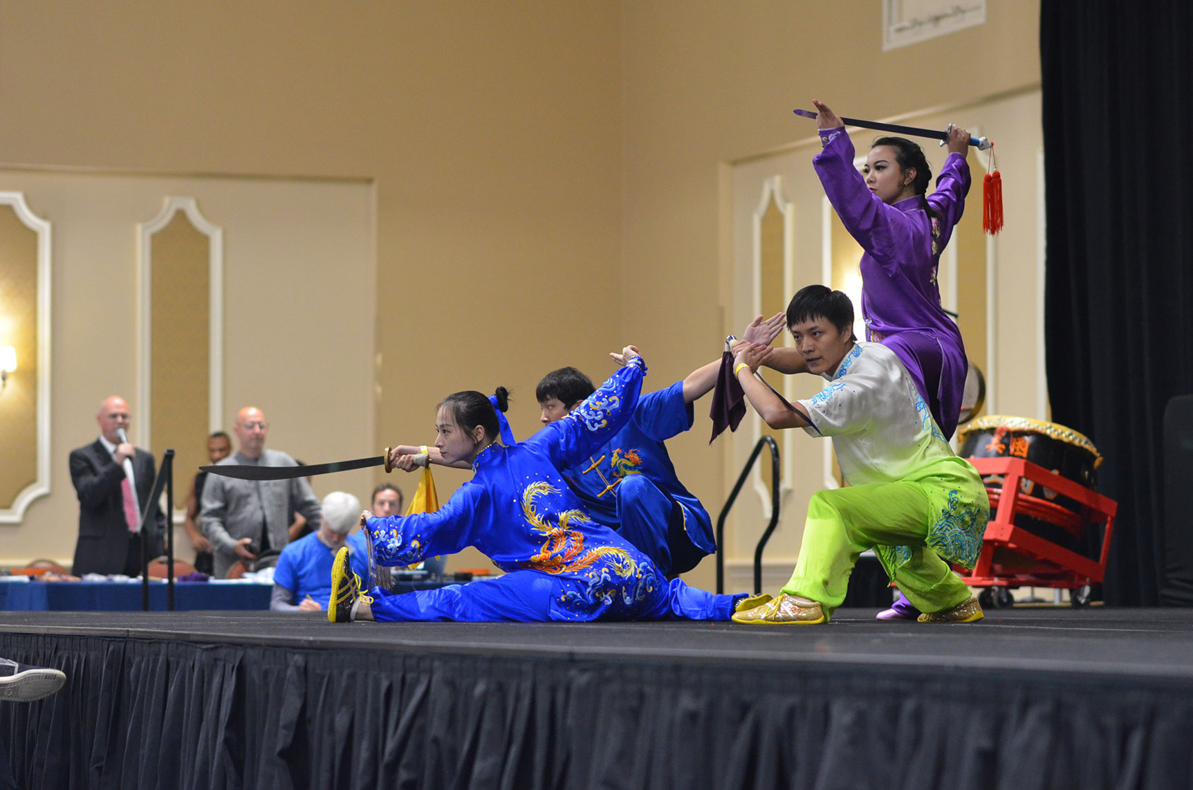 Group of performers in colorful costumes on stage, with some in blue and others in purple, engaging in a dance or martial arts performance.