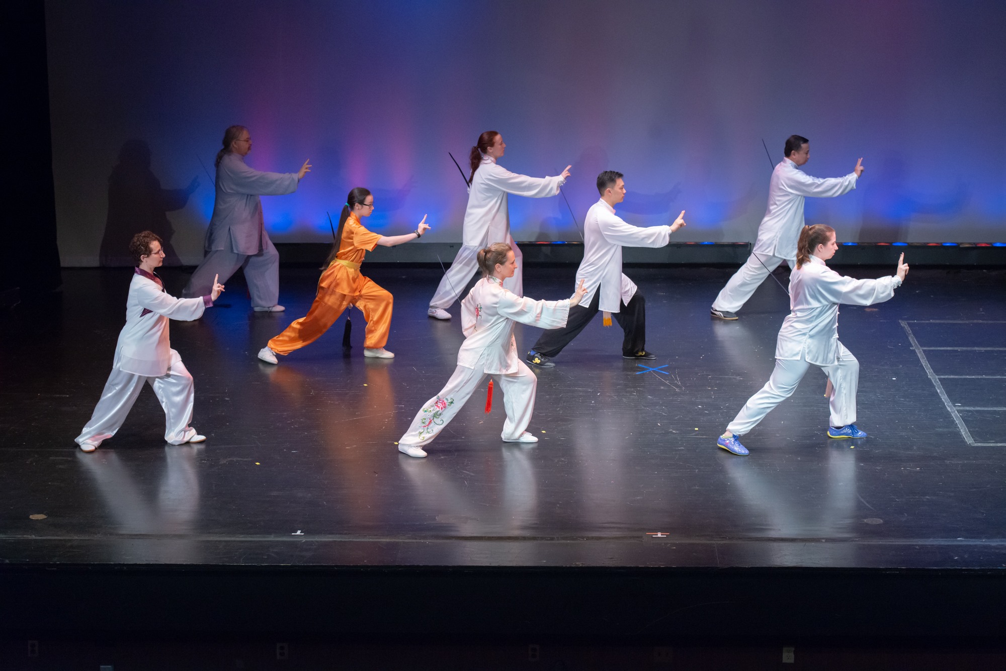 Group of nine people performing a synchronized dance or martial arts routine on stage, wearing white and orange outfits, with a blue gradient background.