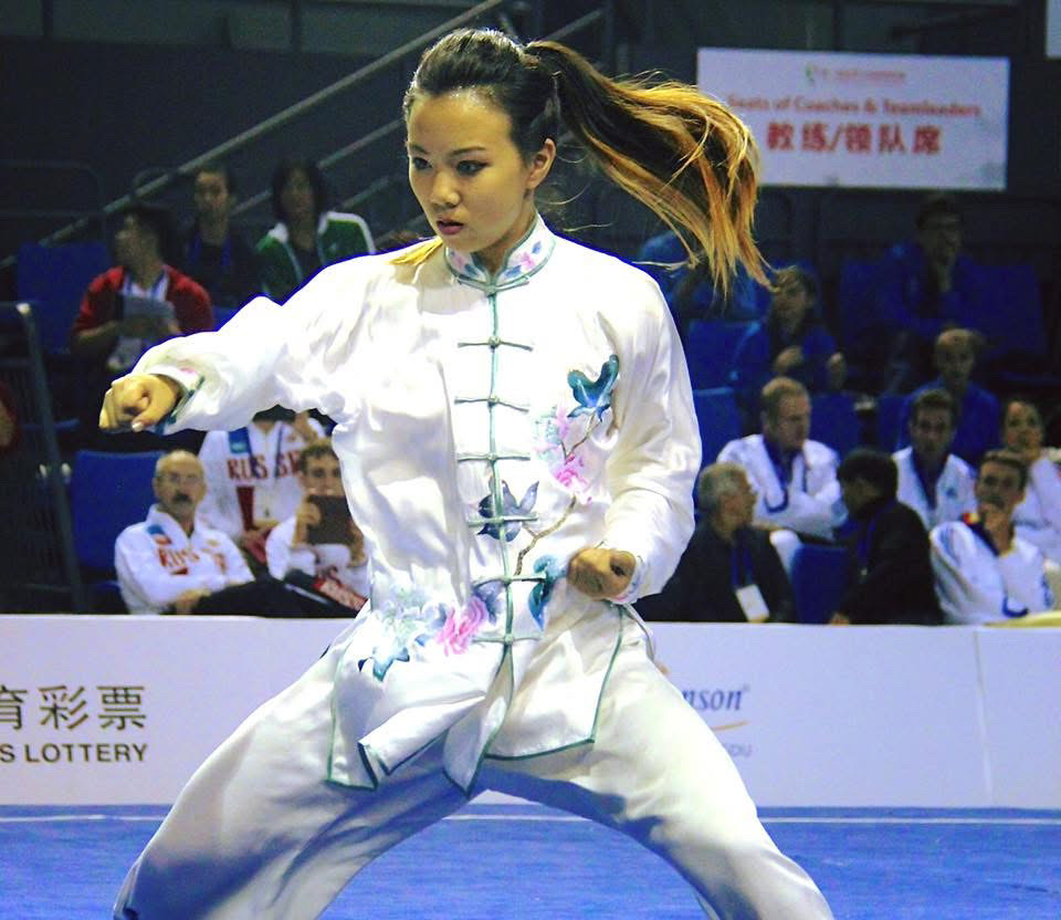 Woman practicing martial arts in a white traditional outfit with colorful embroidery, performing a stance in a sports arena.