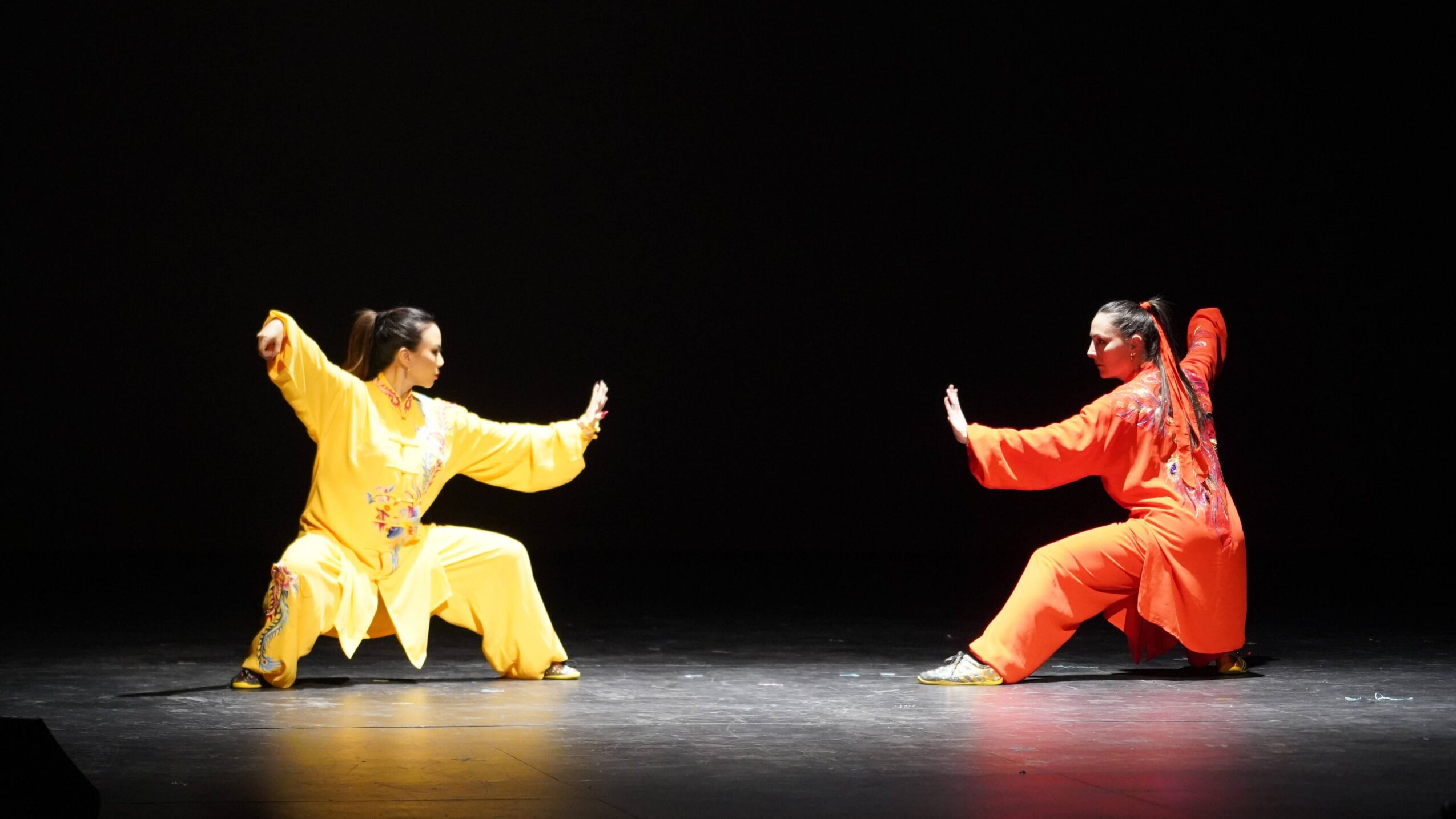Two performers in traditional costumes face each other on stage, one in yellow, the other in red, performing a dance.
