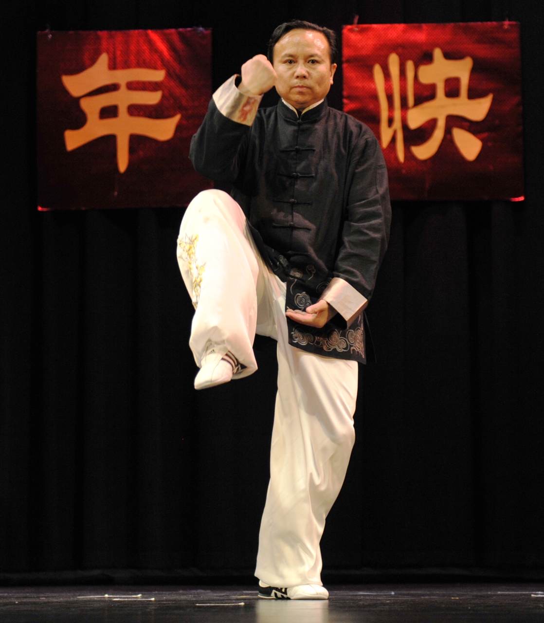 Person performing a martial arts pose on stage with red banners in the background, one with Chinese characters, black and white clothing.