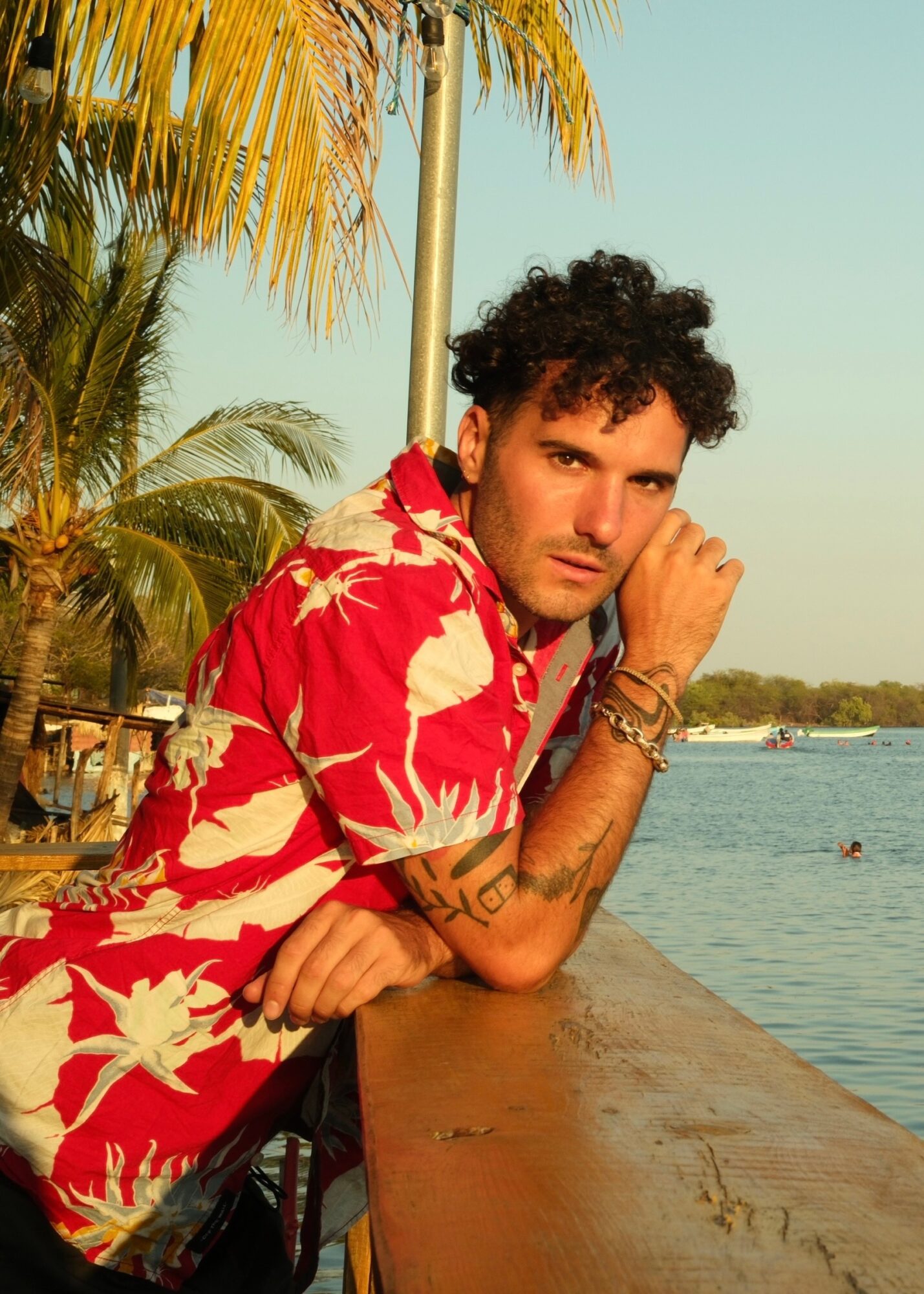Young man with curly hair wearing a red and white floral shirt, resting his chin on his hand by a waterfront.